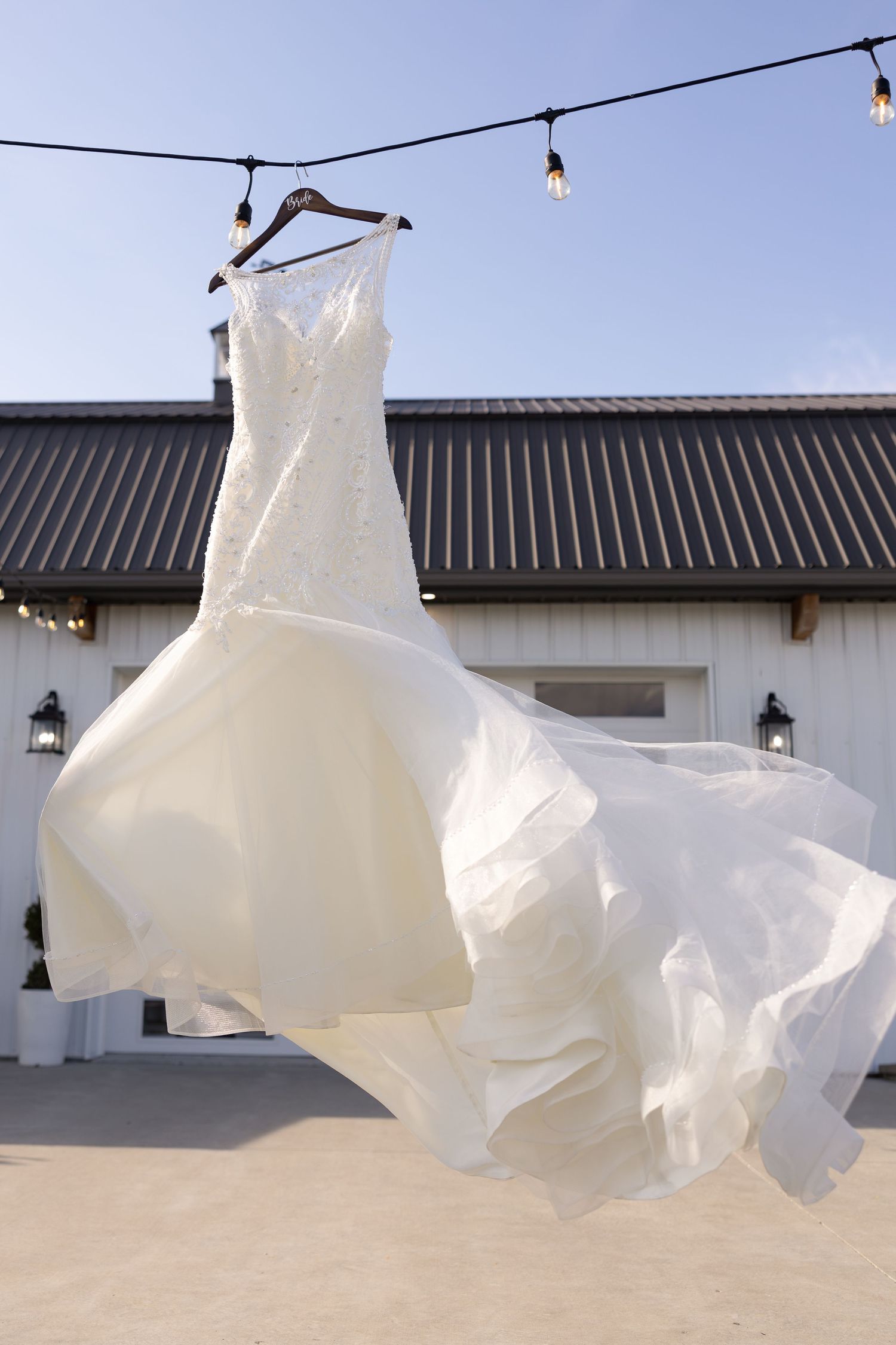 A white wedding dress billows dramatically in the wind while hanging from string lights against a modern venue.