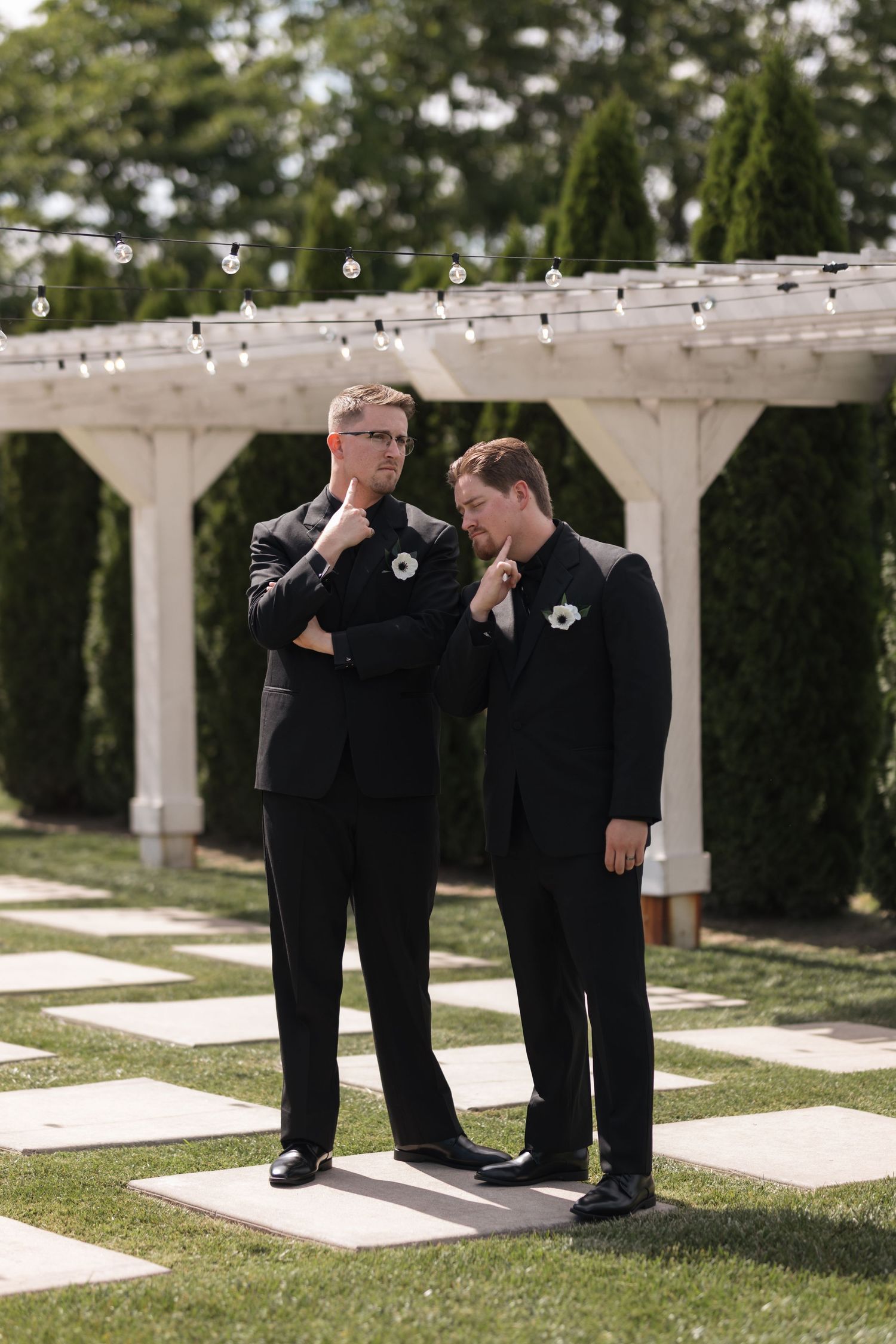 Two people in black suits share an emotional moment under a white pergola with string lights.
