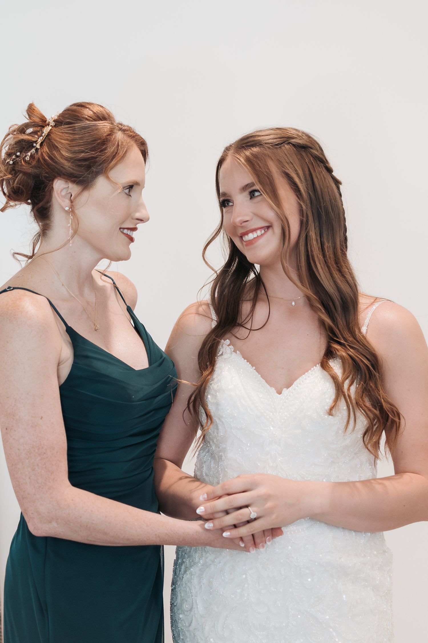 Two women share a joyful moment, one wearing an emerald green dress and the other in a white wedding gown.