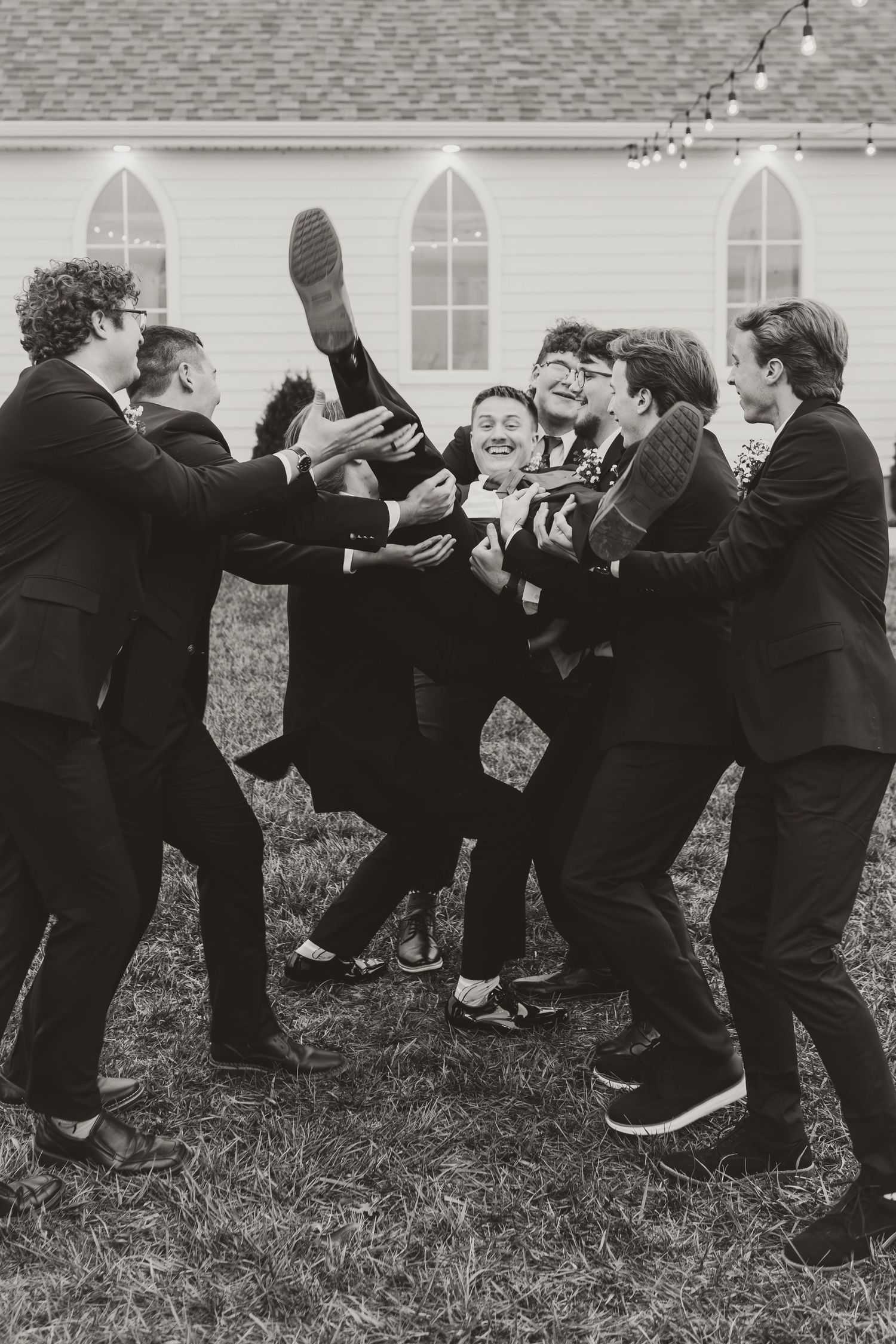 Groomsmen in black suits playfully lift and pose for candid photo outside a church.
