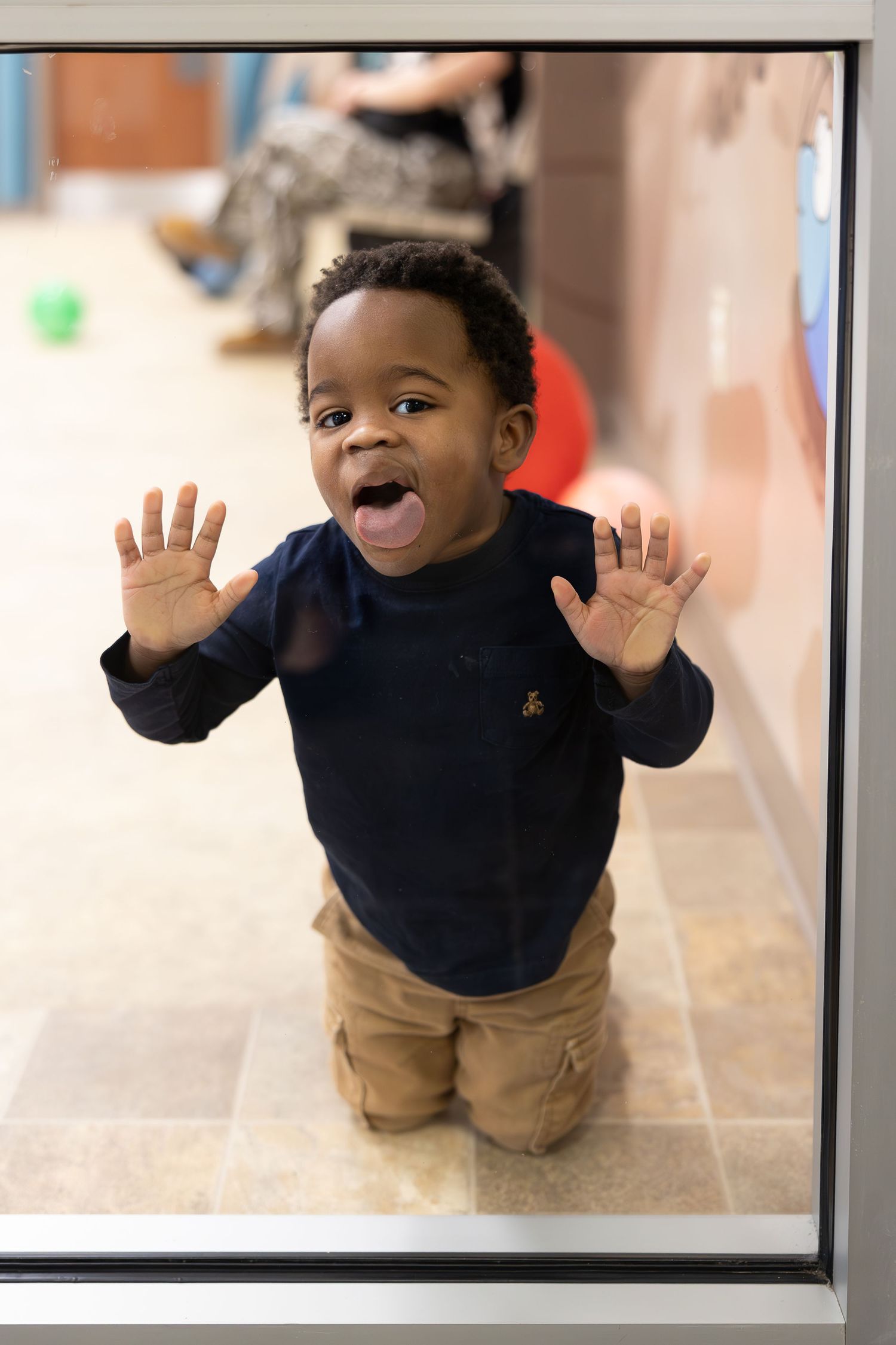 A joyful toddler in navy sweater and khaki pants raises hands while standing behind glass.