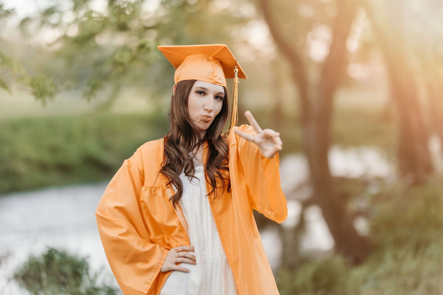 A graduation photo with someone in orange cap and gown posing outdoors by a river.