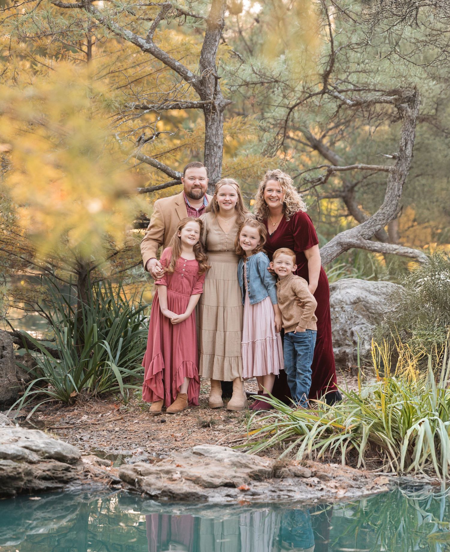 Family portrait in autumn setting by a reflective pond with colorful fall foliage in the background.