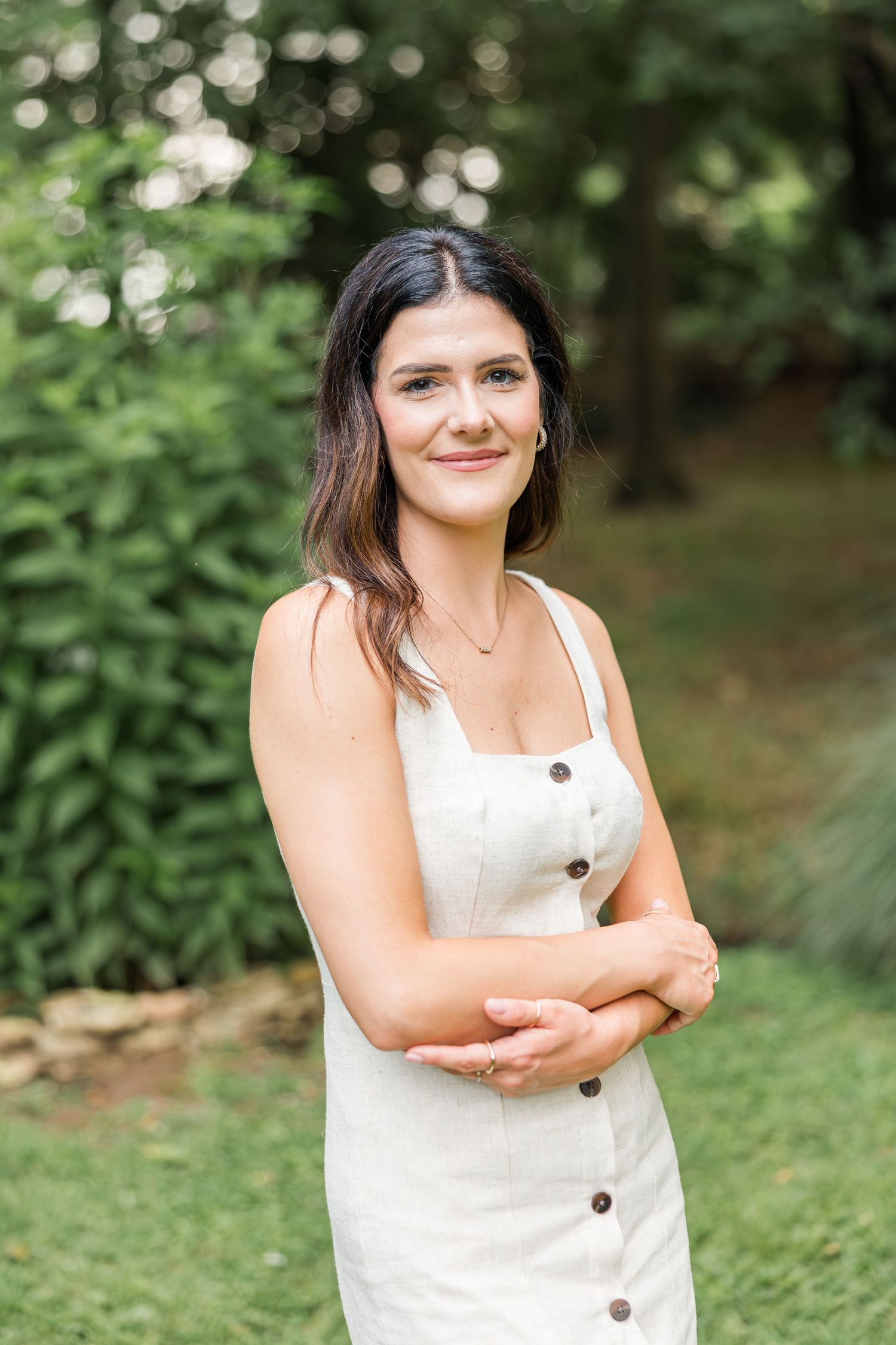 A person in a white summer dress with buttons stands outdoors against a green natural background.