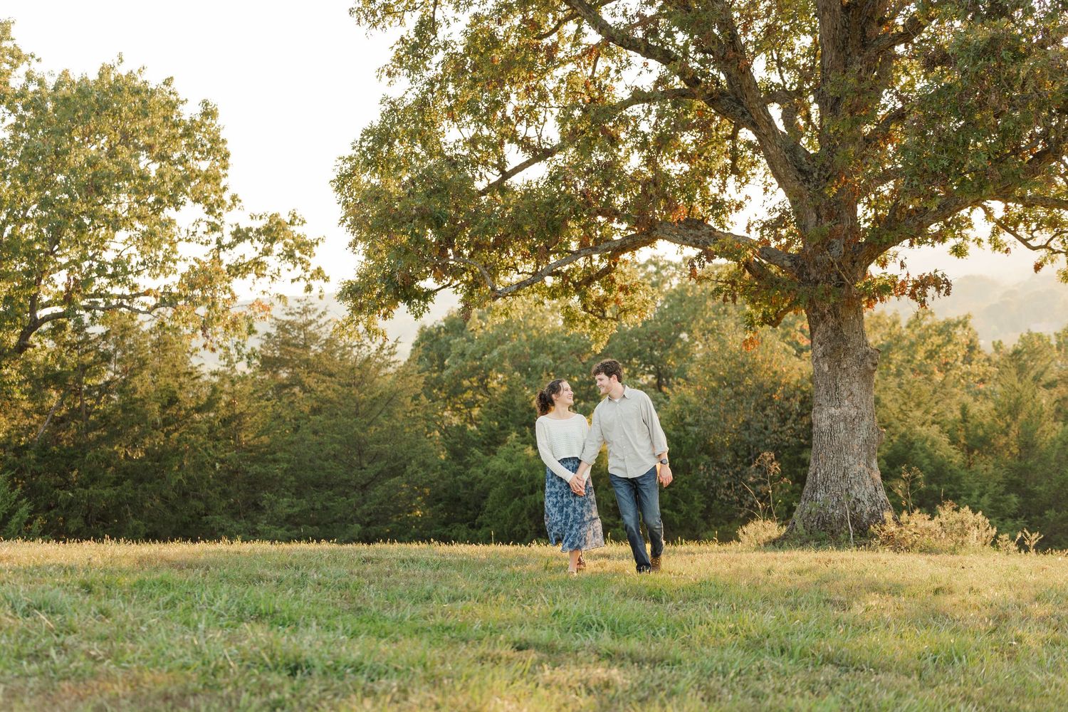 A romantic couple walks together through a sunlit meadow during golden hour at sunset.