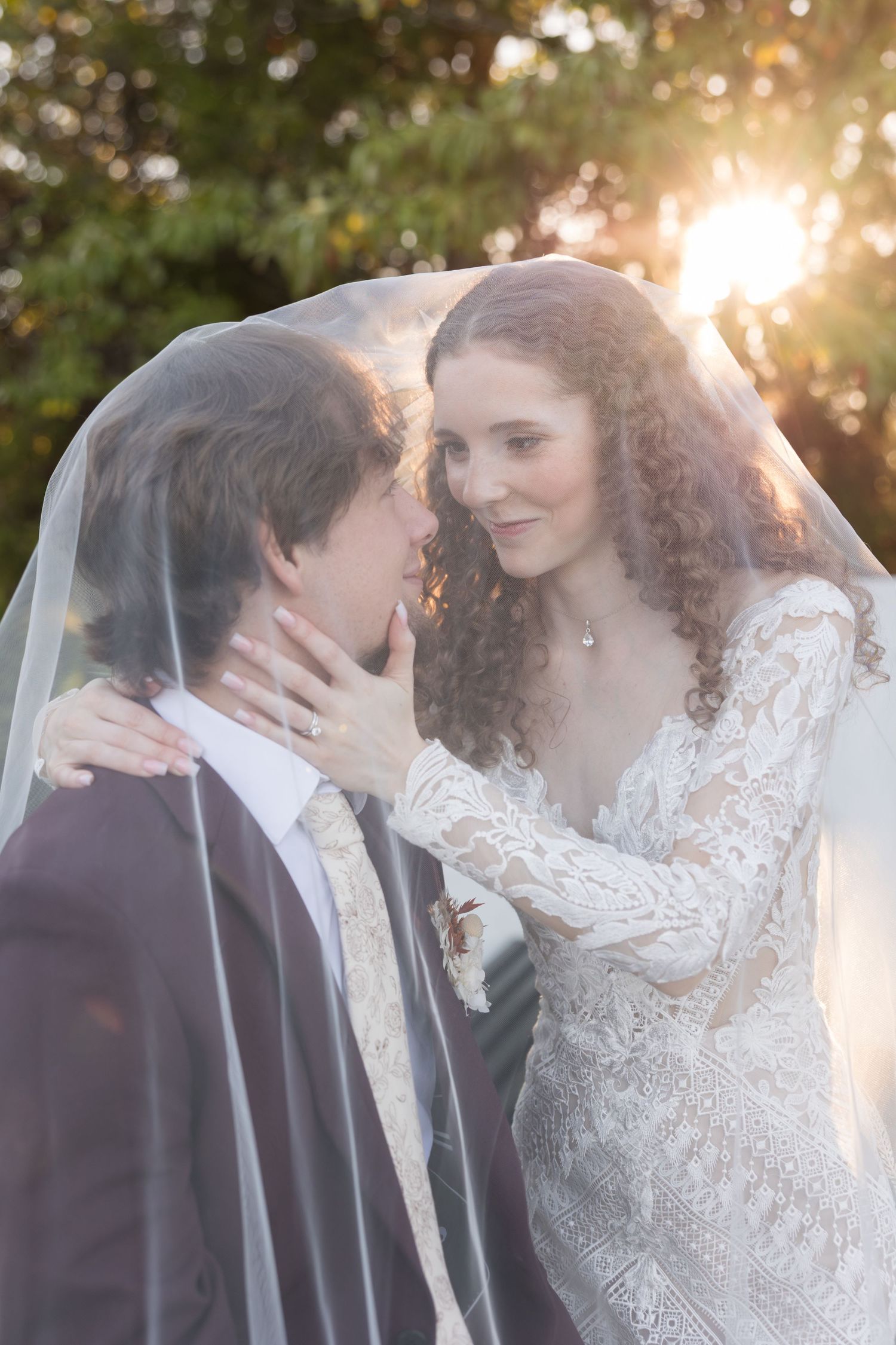 A bride and groom share an intimate moment under a veil with golden sunset light glowing behind them.
