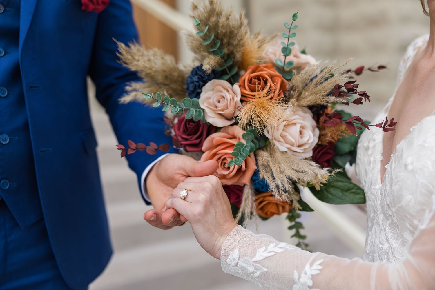 Bride and groom holding hands with a vibrant boho bouquet of orange, pink, and burgundy roses with pampas grass.