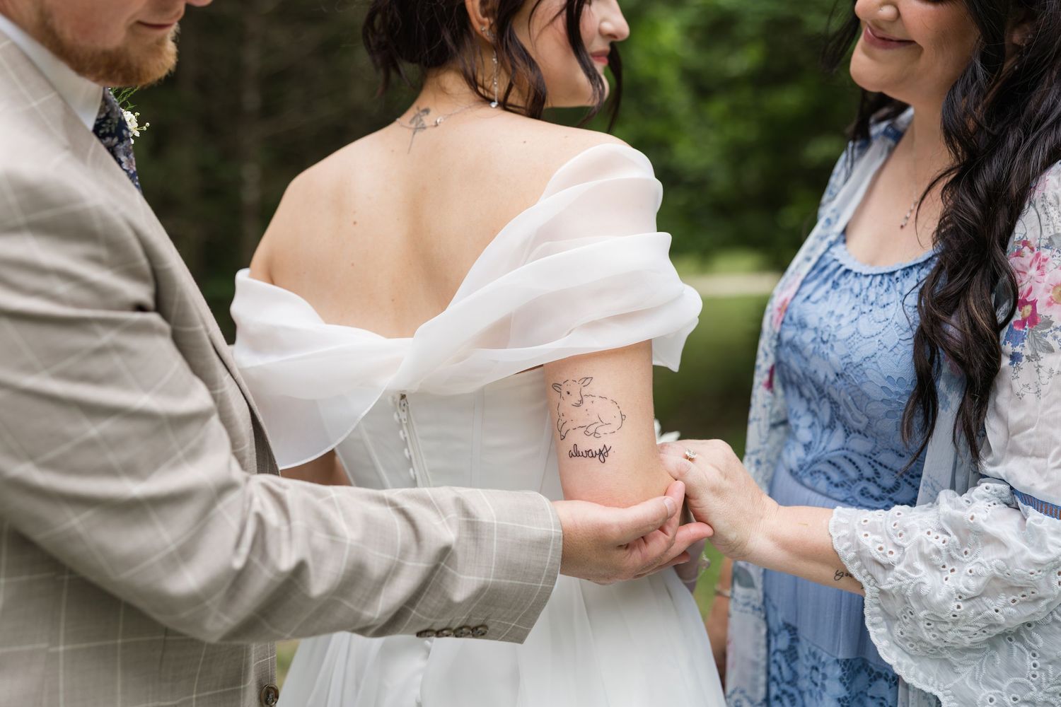 Bride with tattoo on arm in off-shoulder wedding dress being helped by groom and bridesmaid outdoors.