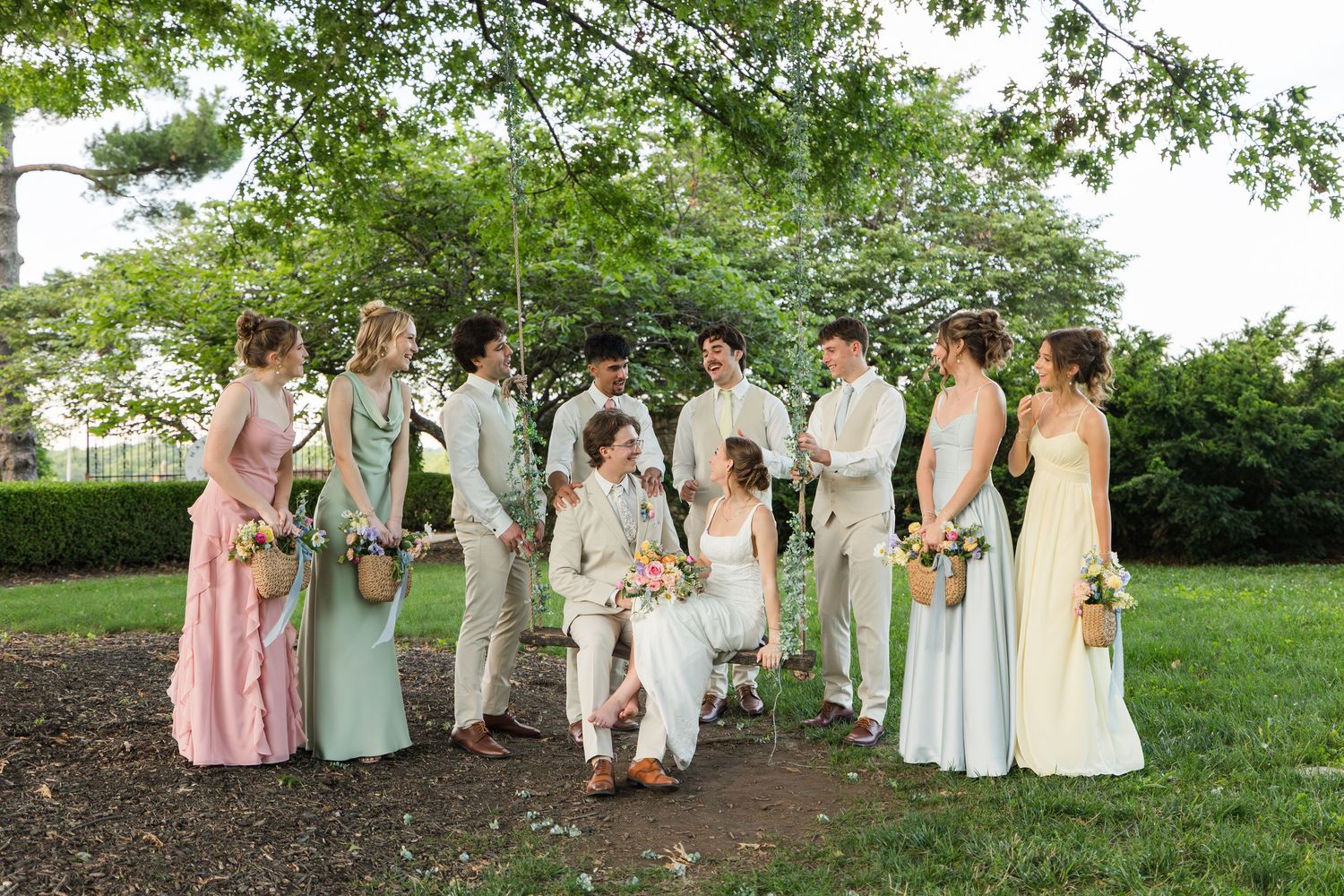 Wedding party in pastel gowns and tan suits gathered outdoors under lush green trees with bride and groom centered.