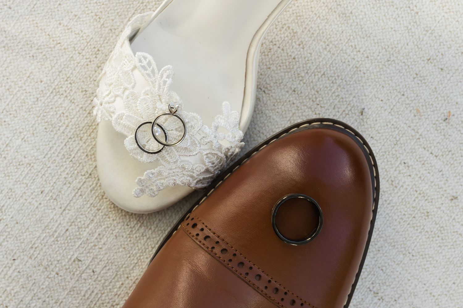Wedding rings resting on a white lace bridal shoe and brown leather Oxford dress shoe on a linen background.