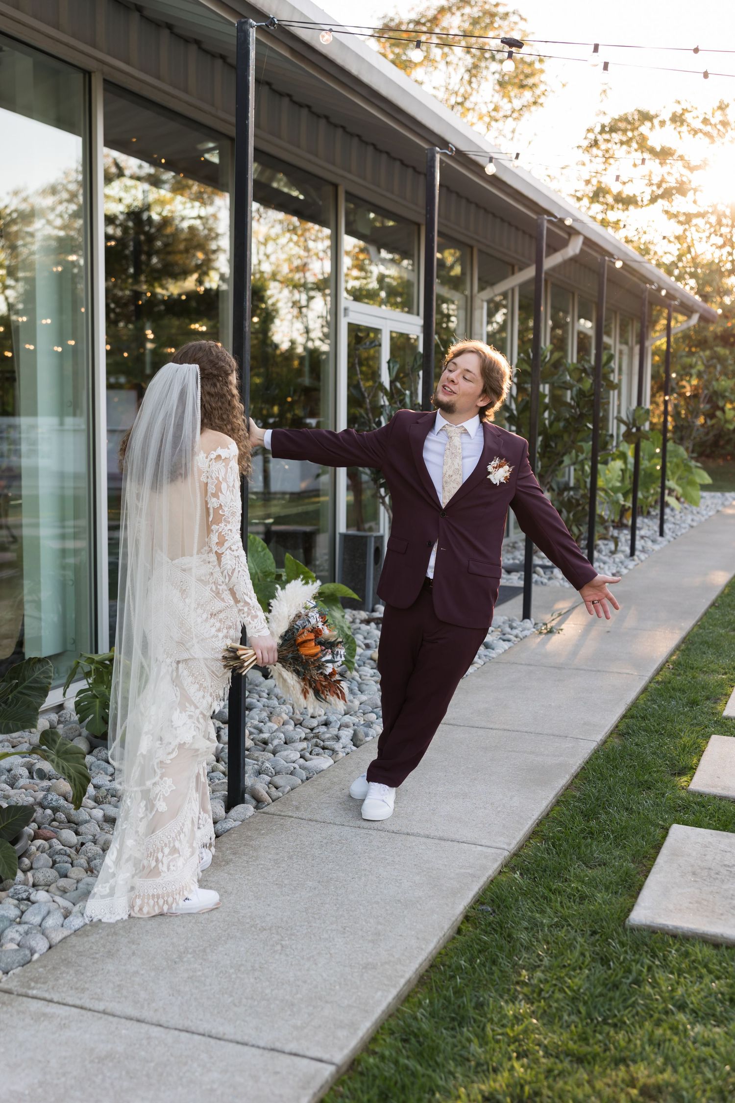 Groom in burgundy suit dramatically gestures toward bride in lace gown outside glass venue building.