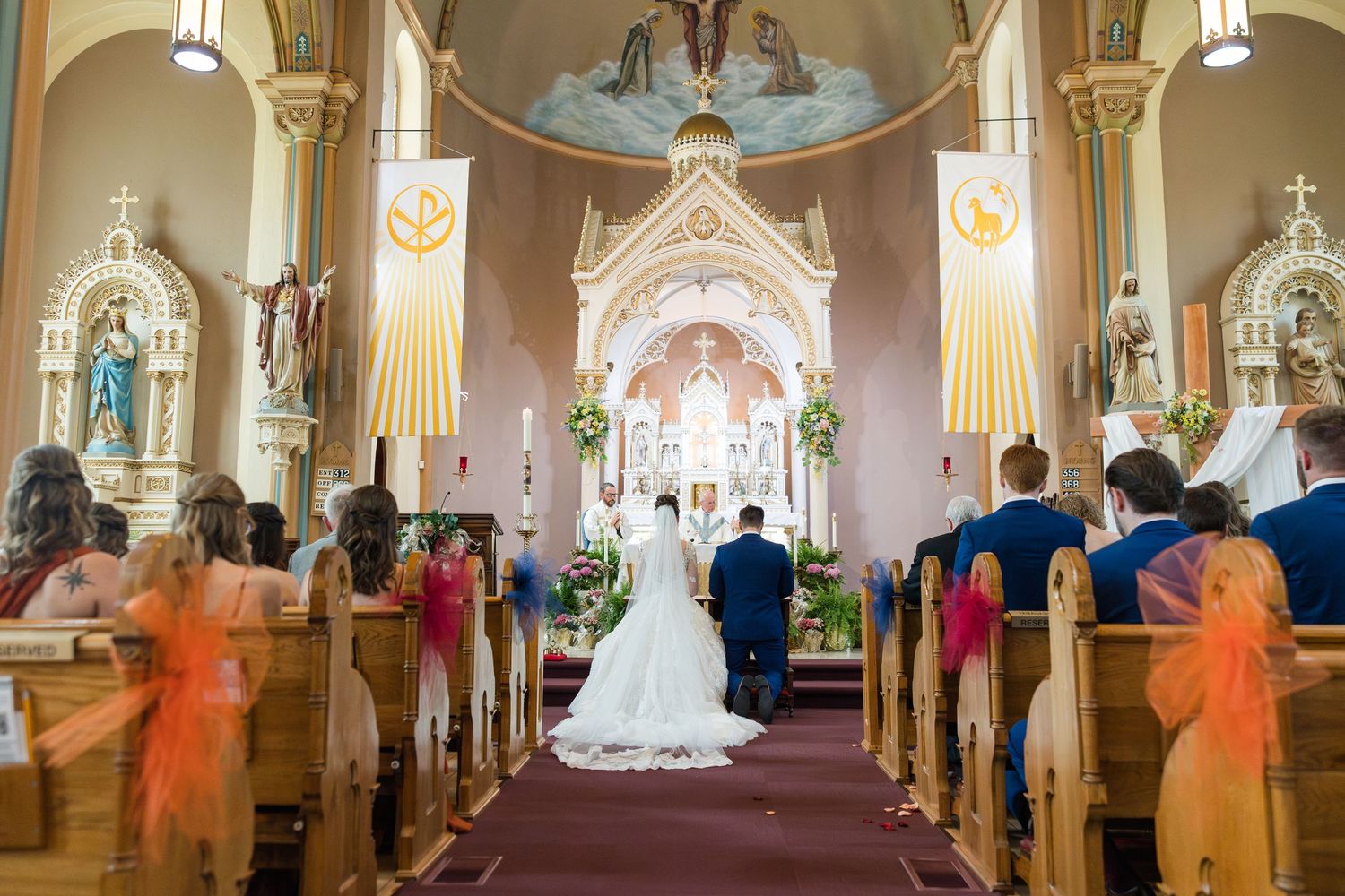 A bride and groom stand at the altar of an ornate Catholic church surrounded by guests seated in wooden pews.