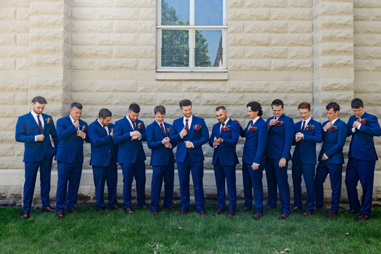 Groomsmen in matching navy blue suits adjust their ties and boutonnieres outside a stone building.