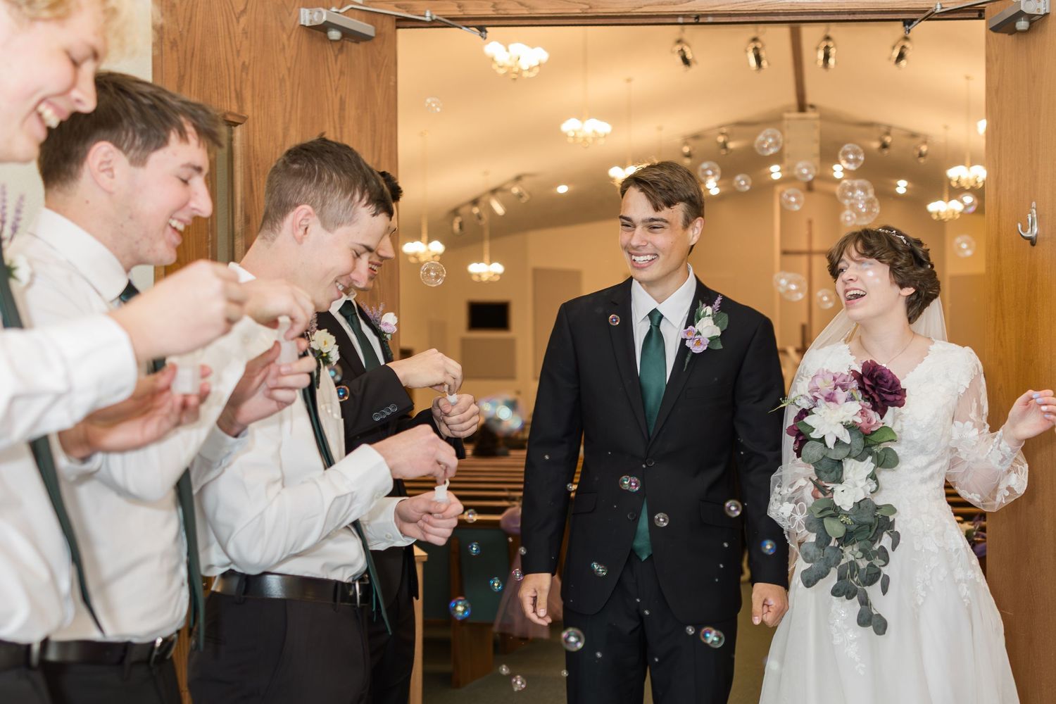 Wedding party celebrates with bubbles during indoor reception as couple smiles joyfully in formal attire.