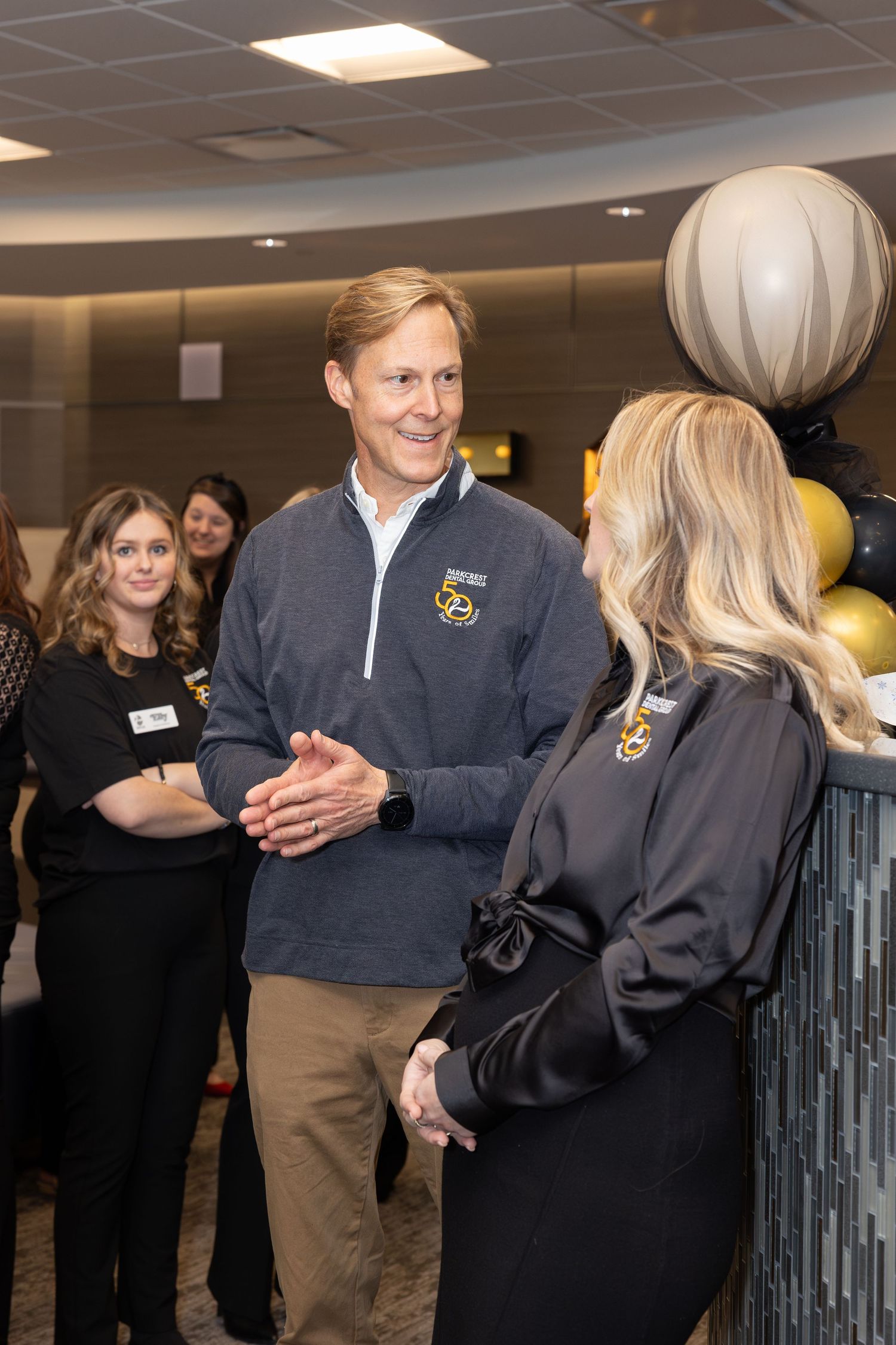 People in branded company attire interact during an office celebration with gold balloons.