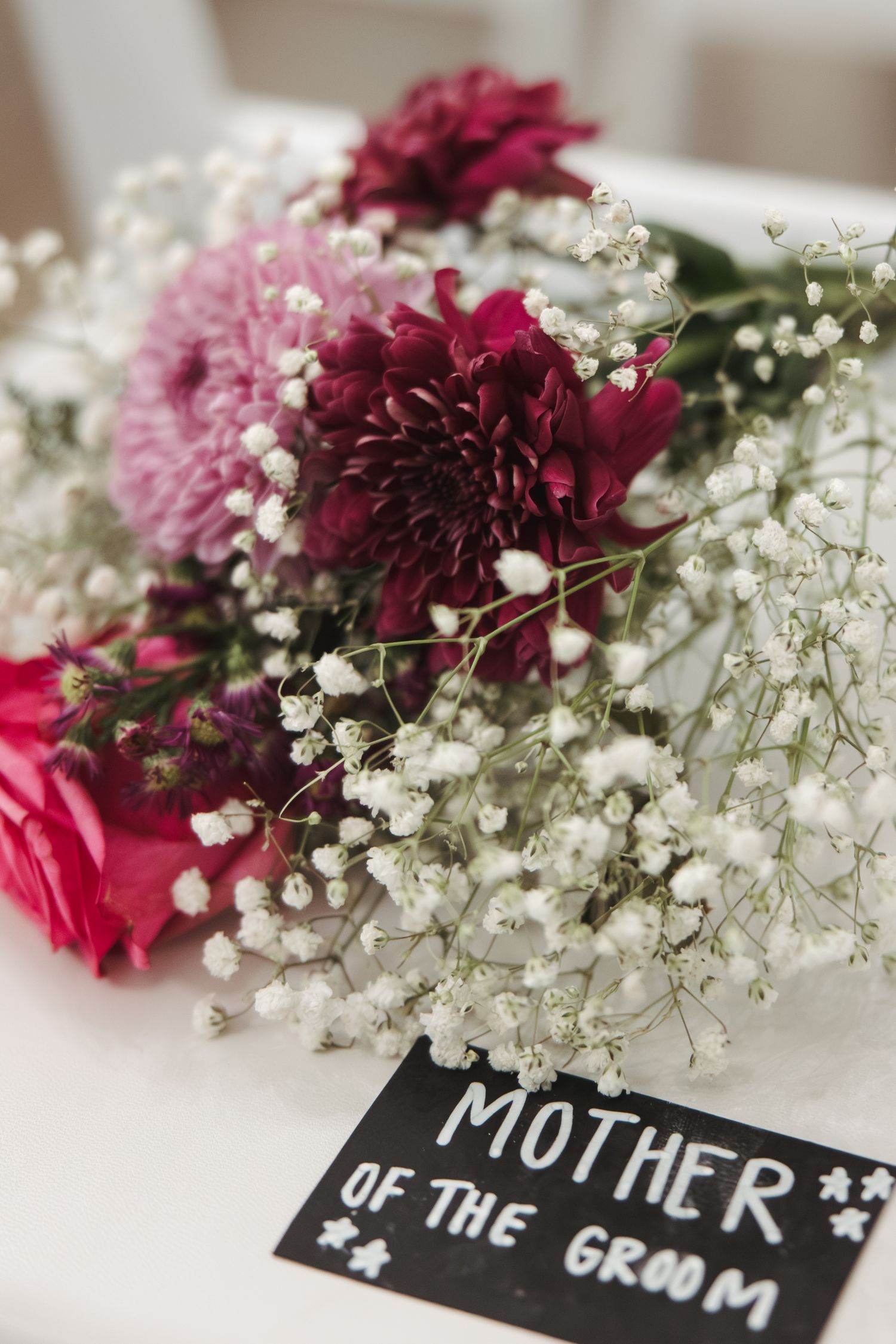 A floral arrangement with pink and burgundy flowers alongside baby's breath next to a 'Mother of the Groom' sign.