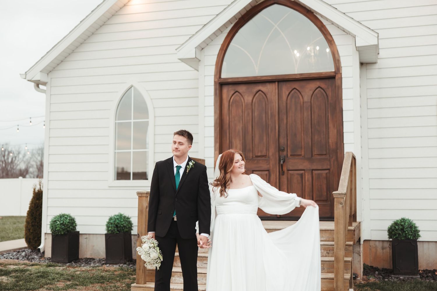 Wedding couple poses in front of a charming white chapel with wooden doors and topiary.