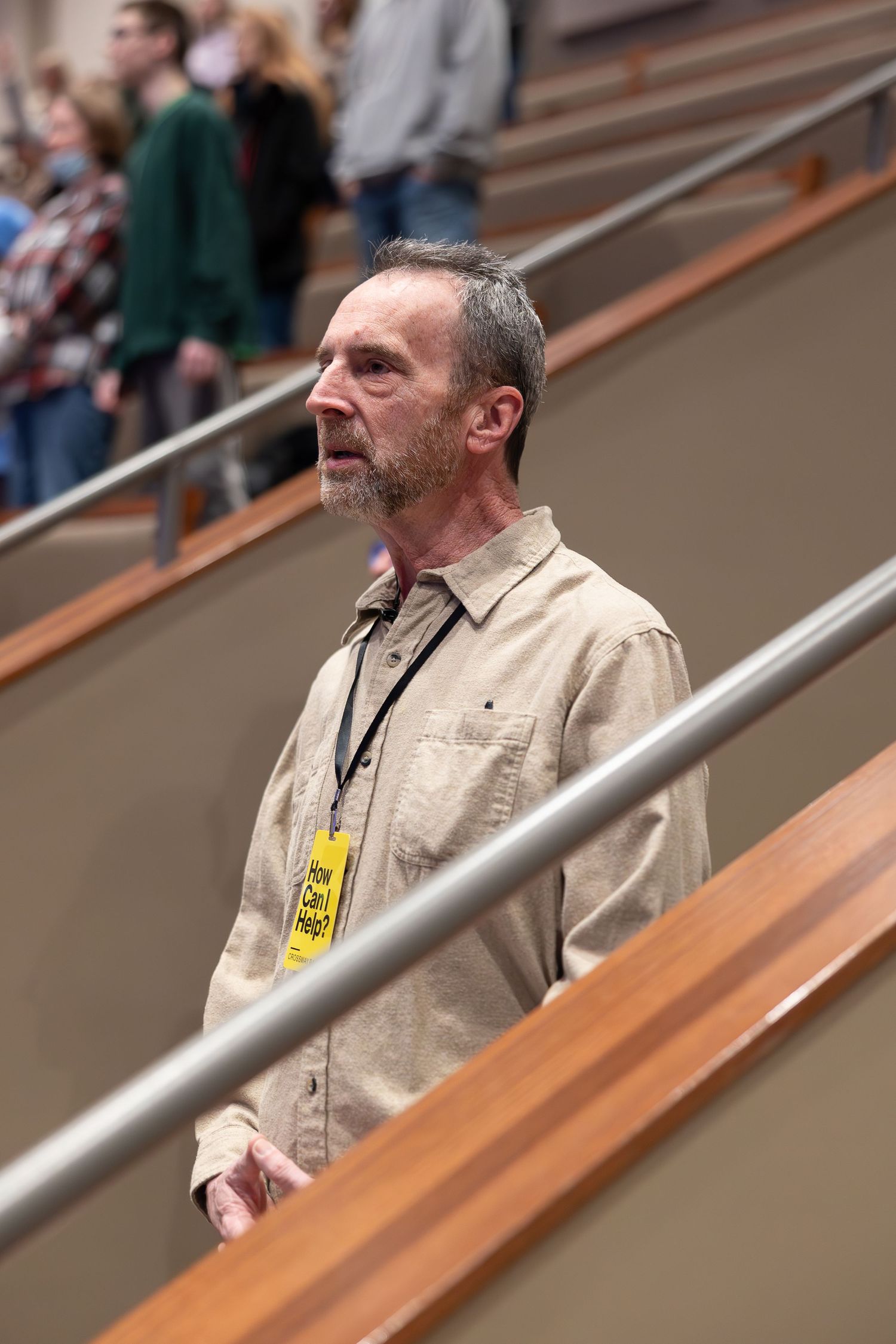 Attendee standing in tiered seating area wearing name badge and tan shirt.