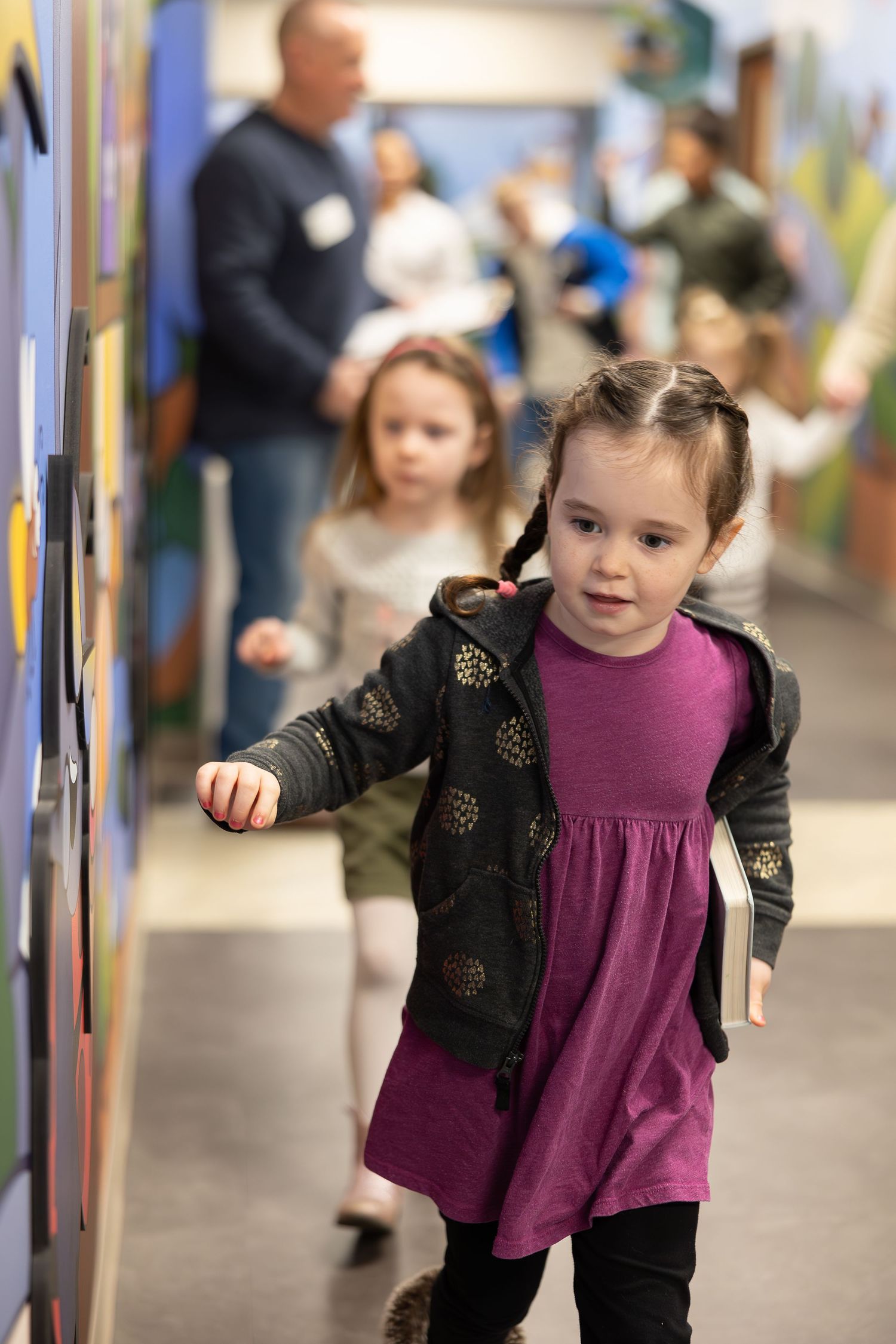 Child in purple dress and black sweater walking along colorful hallway wall.