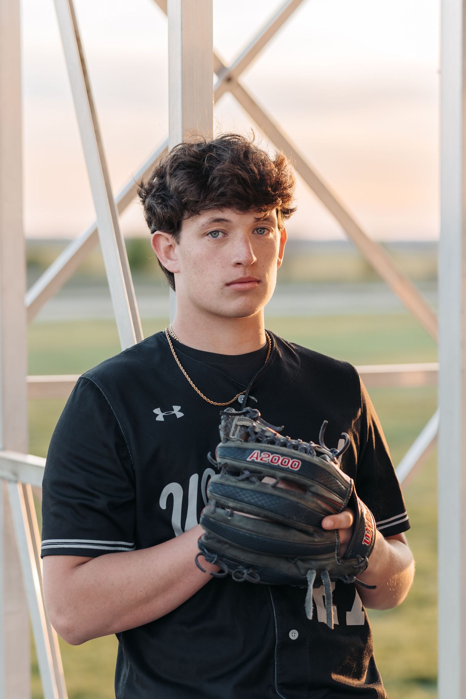 Baseball player in dark uniform holds glove while posing against metal structure at sunset.