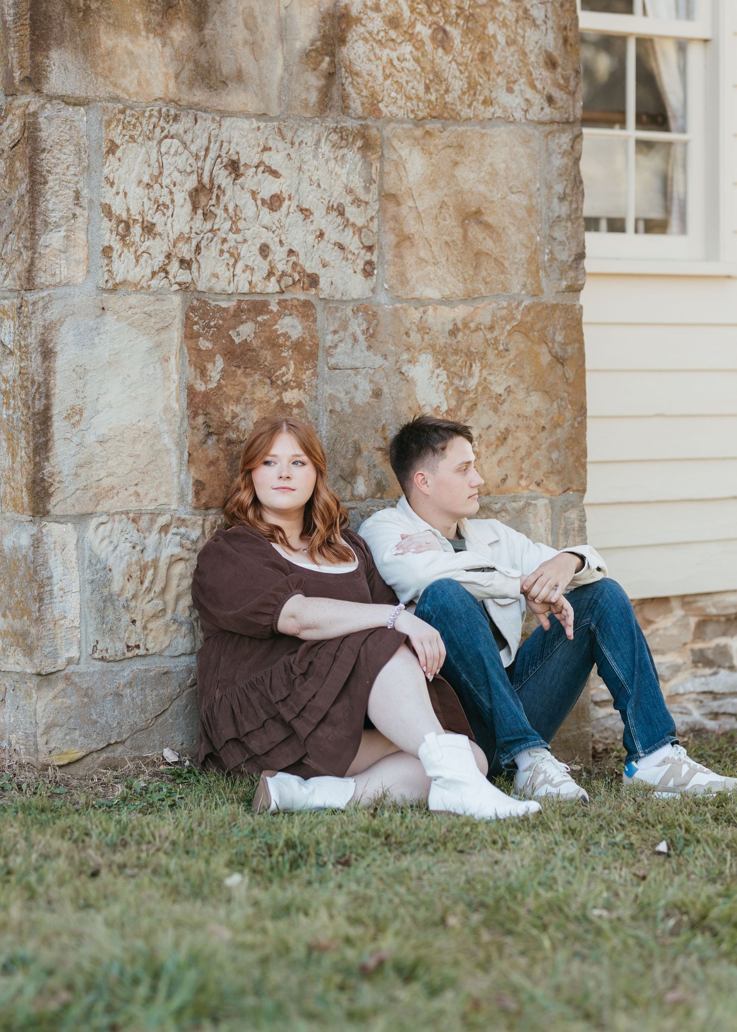 Two people rest against a weathered stone wall on a grassy lawn.
