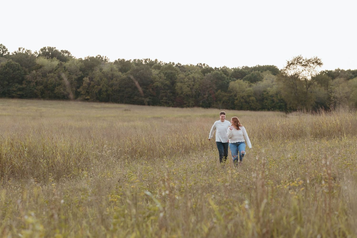 Couple walking together through a rustic field with autumn trees in the distance.