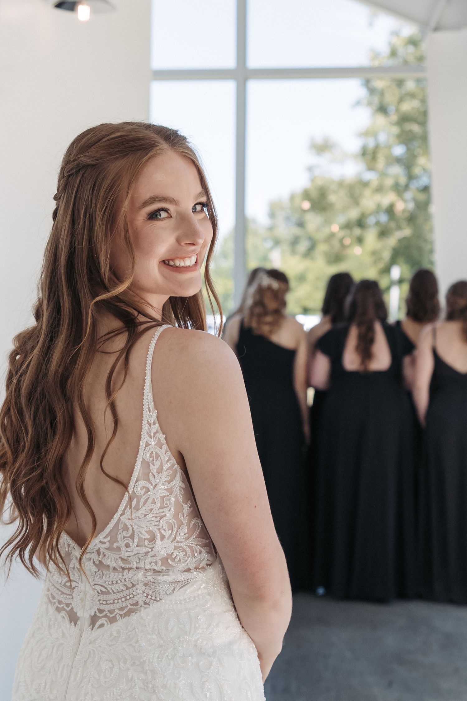 Elegant bride in lace dress smiling over shoulder with bridesmaids in background.