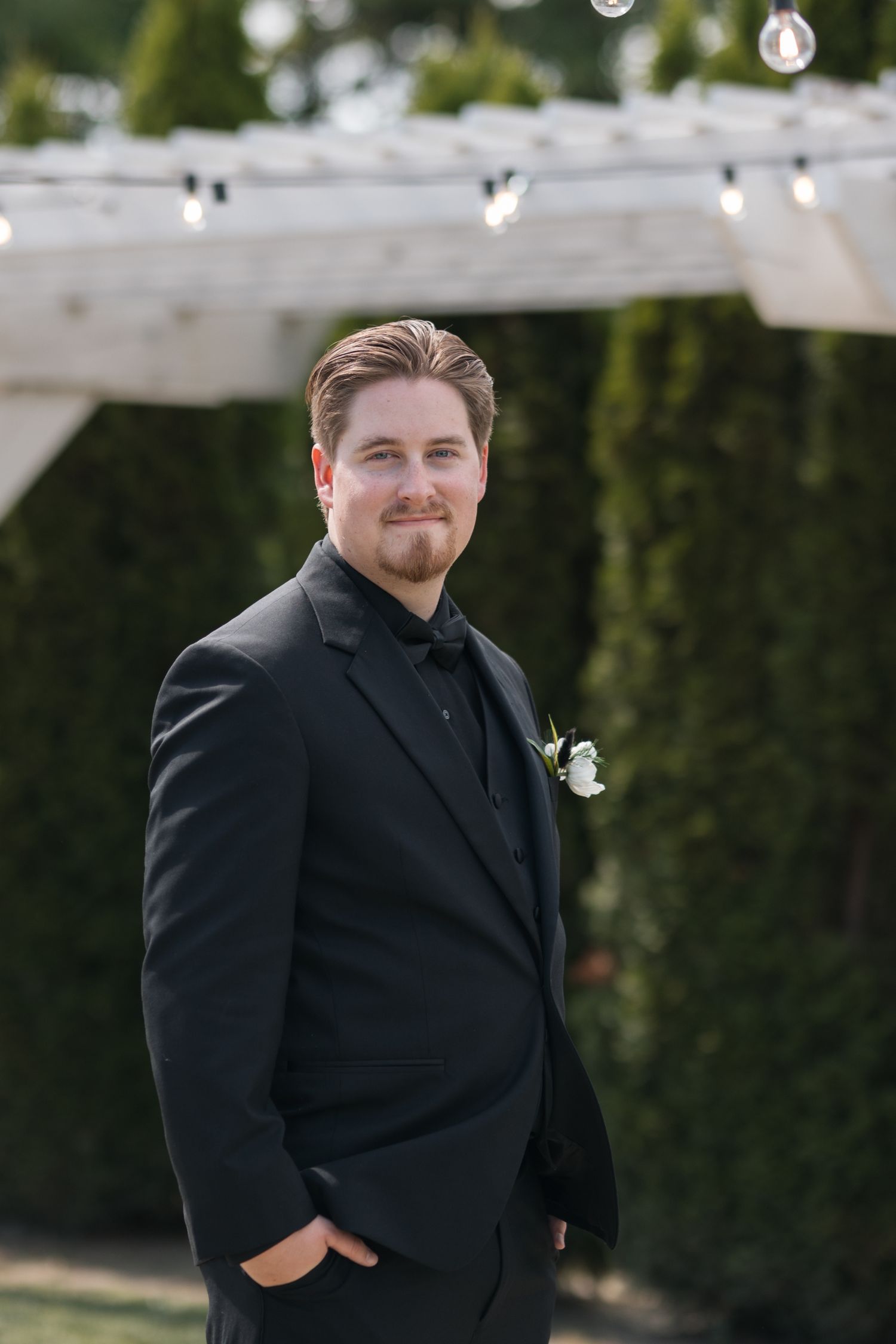 Groom in classic black tuxedo poses under string lights at outdoor wedding venue.