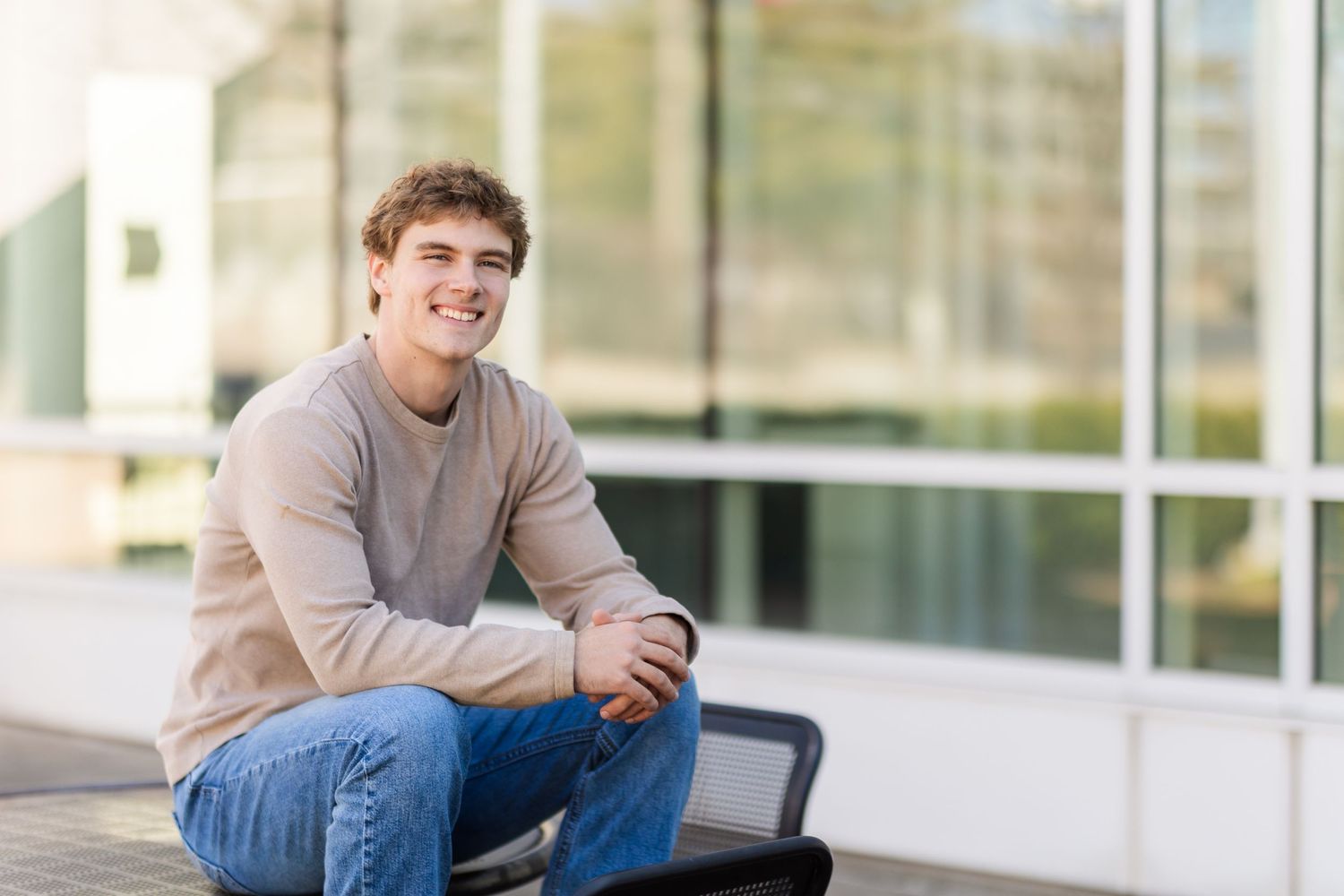 Person in tan sweater sits casually outside modern glass building.