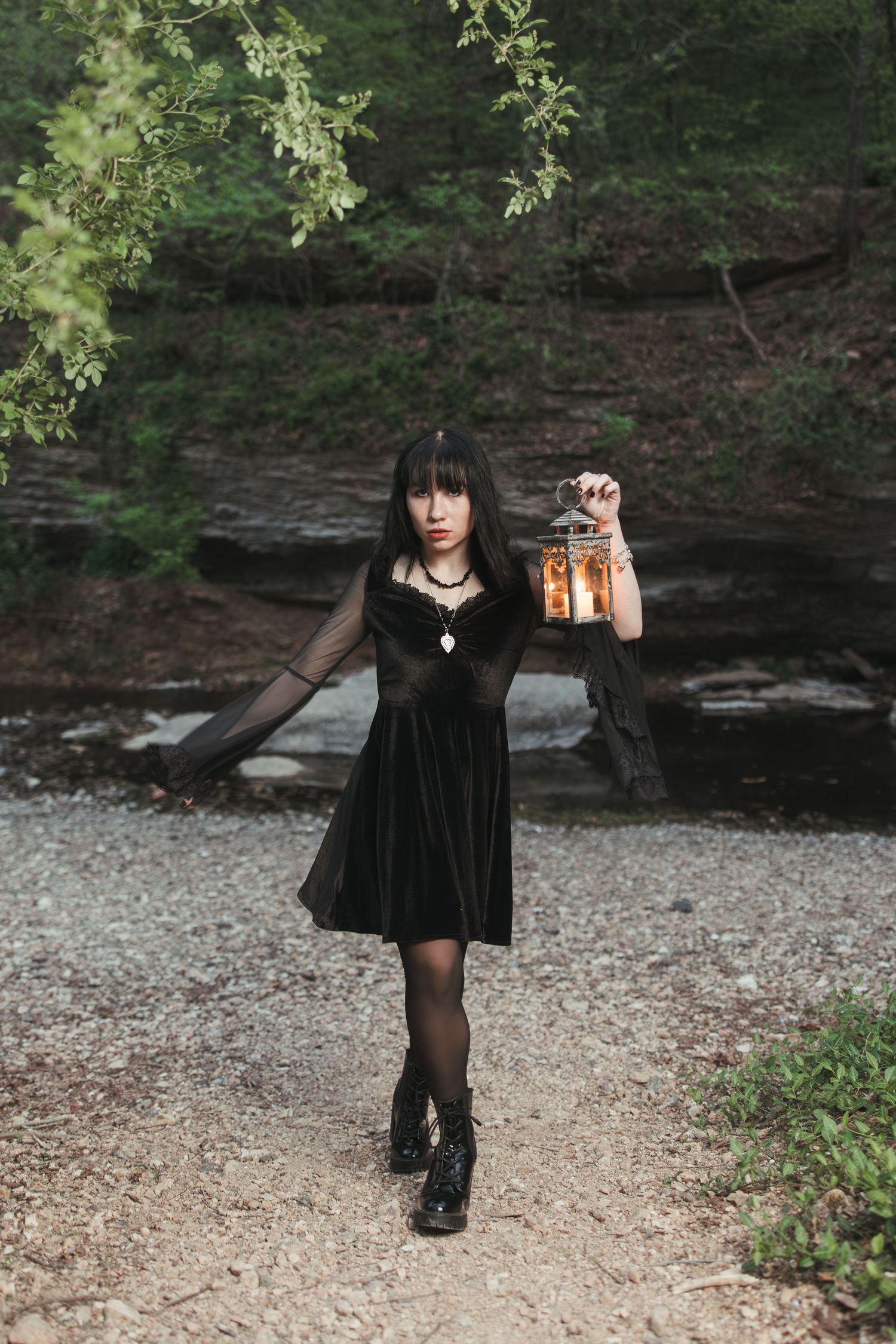 A person in a black velvet dress holds up a vintage lantern while standing on a forest path.
