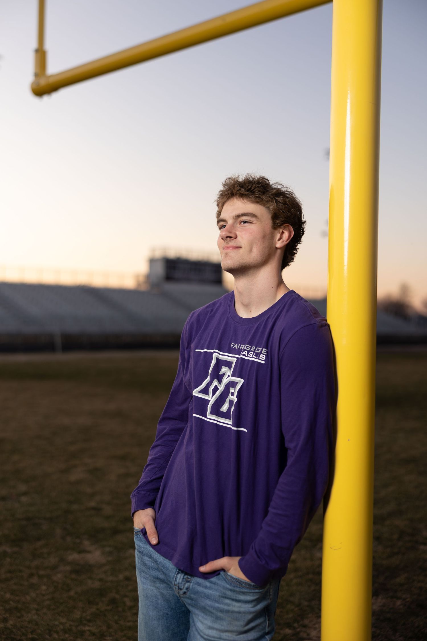 Athletic scene with purple school spirit shirt posed by yellow goal post at sunset.