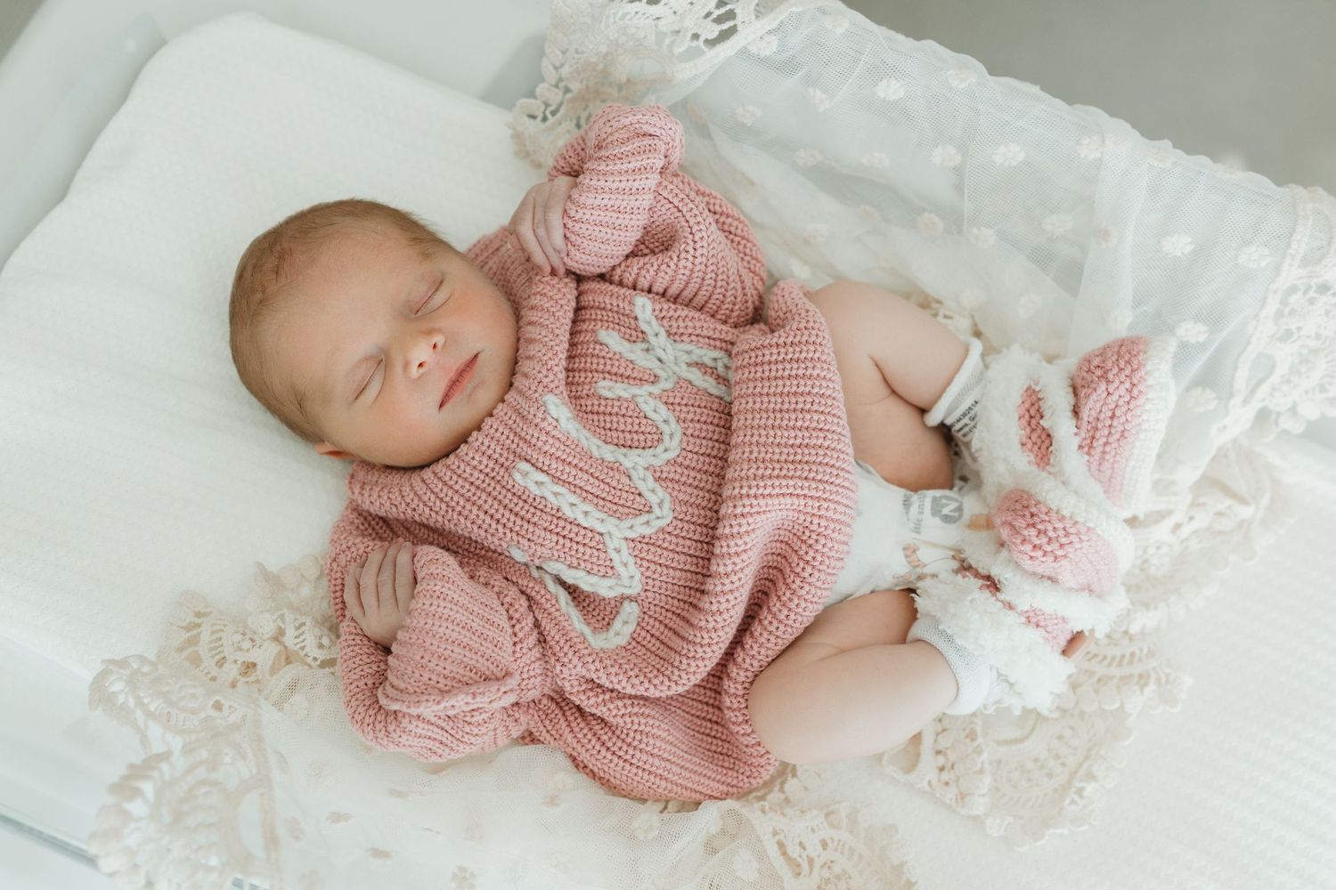 A sleeping newborn wearing a soft pink knit sweater lies on white lace bedding.