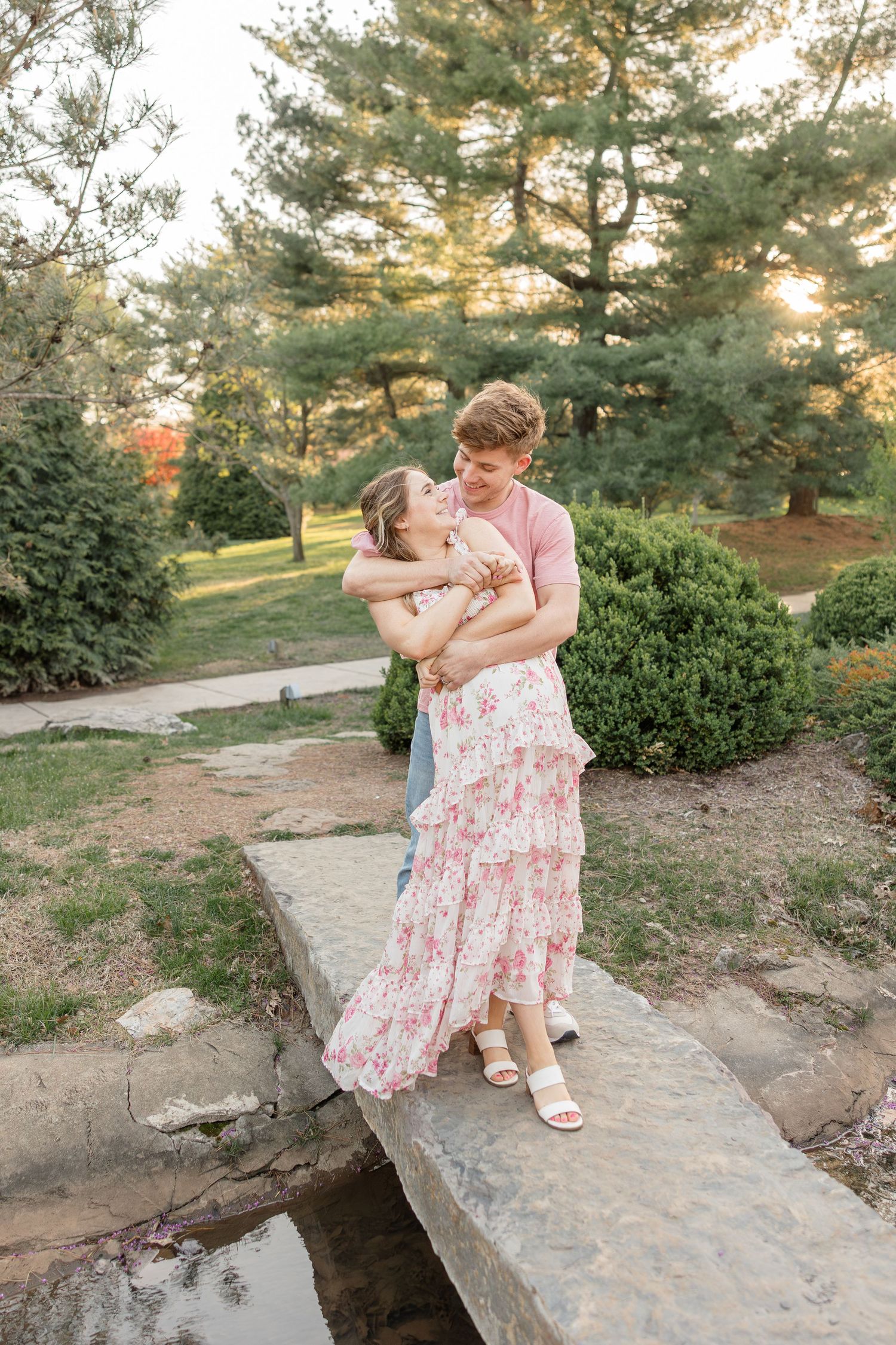 A couple embraces on a stone bridge path surrounded by pine trees at sunset.