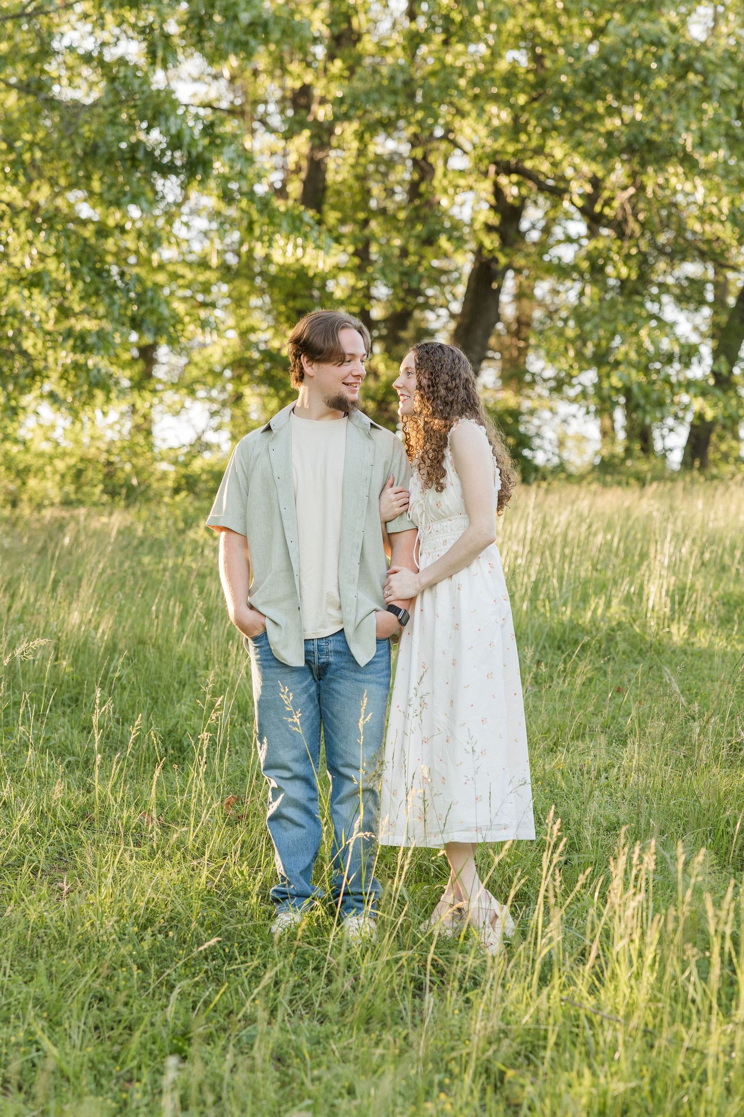 A couple stands together in a sunlit meadow surrounded by trees on a warm summer evening.