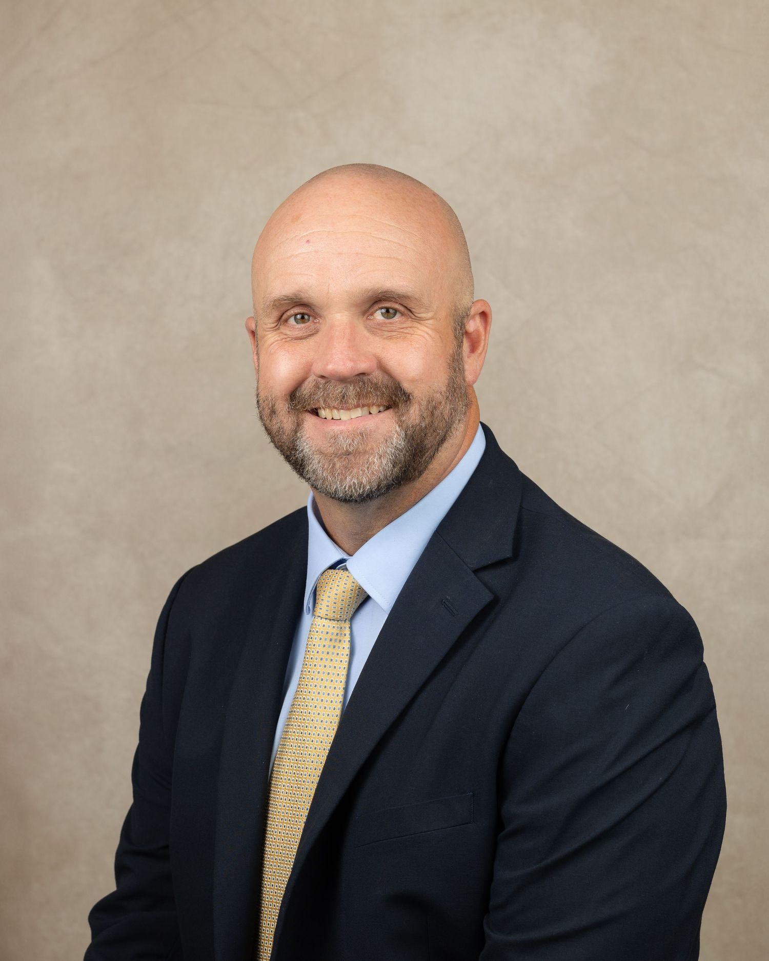 Professional headshot of person in dark business suit with yellow tie against neutral backdrop.