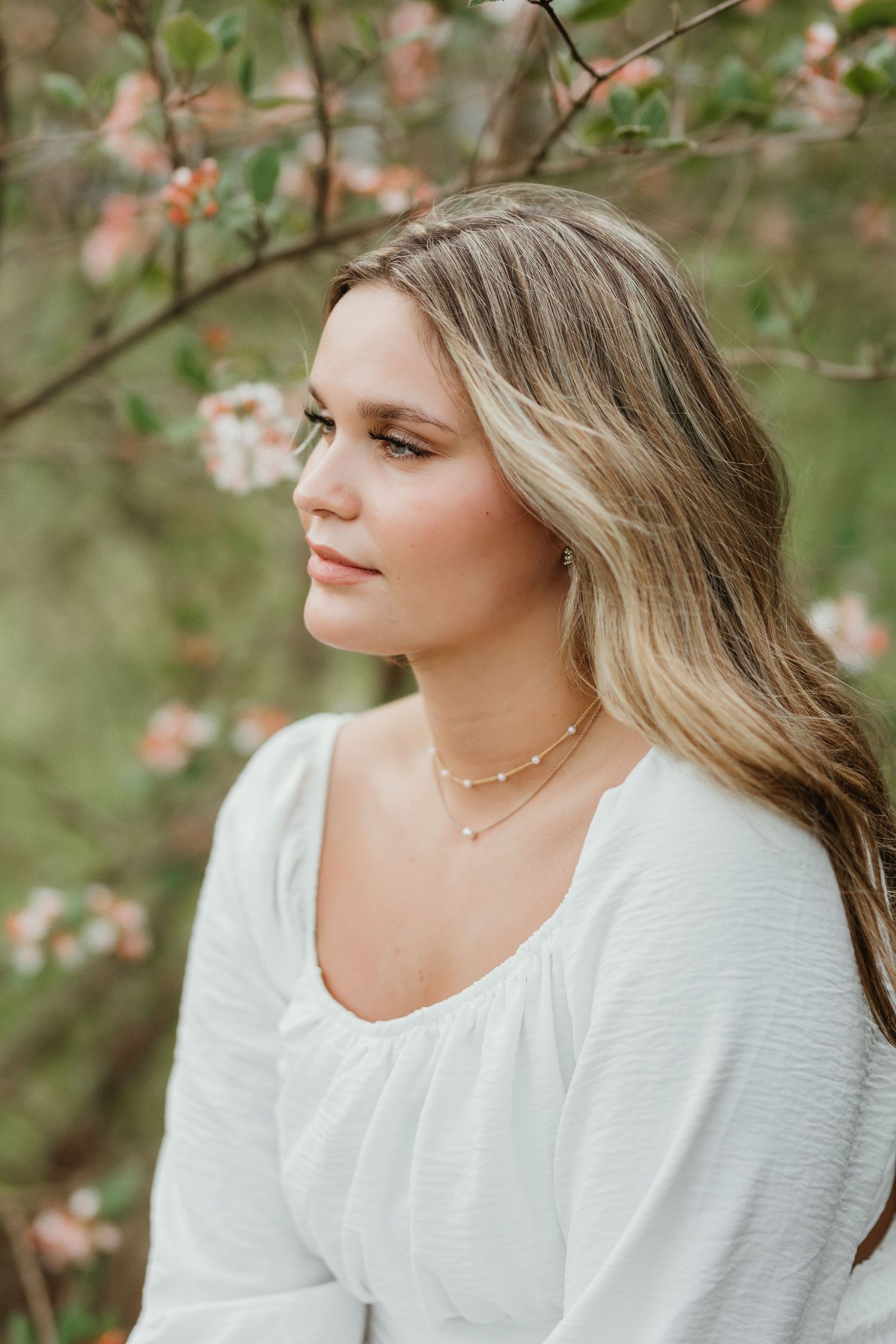 Portrait in a white blouse with delicate necklace posed among soft pink flowering branches in spring.
