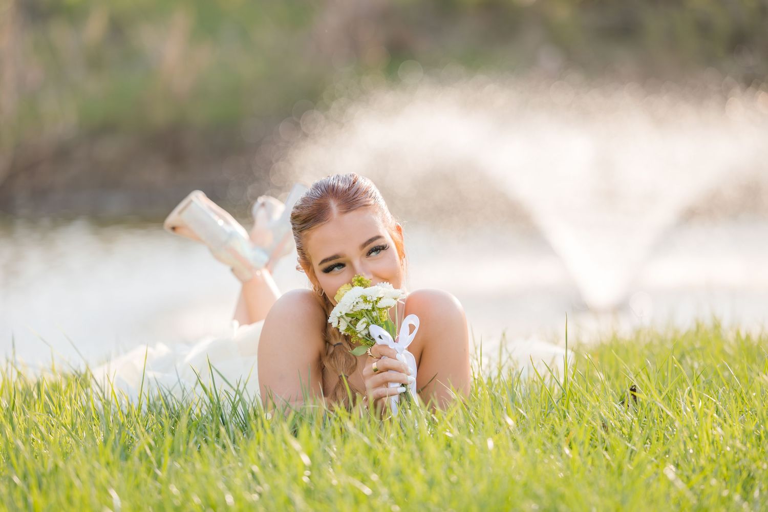 A person lies in green grass holding wildflowers on a sunny day with a fountain spraying water in the background.