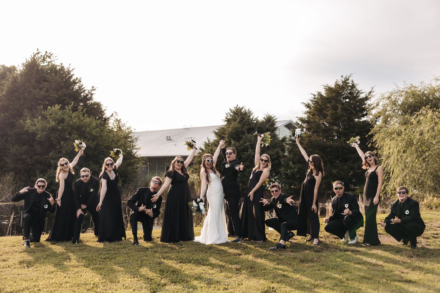 Wedding party in black attire poses enthusiastically on grassy hillside.