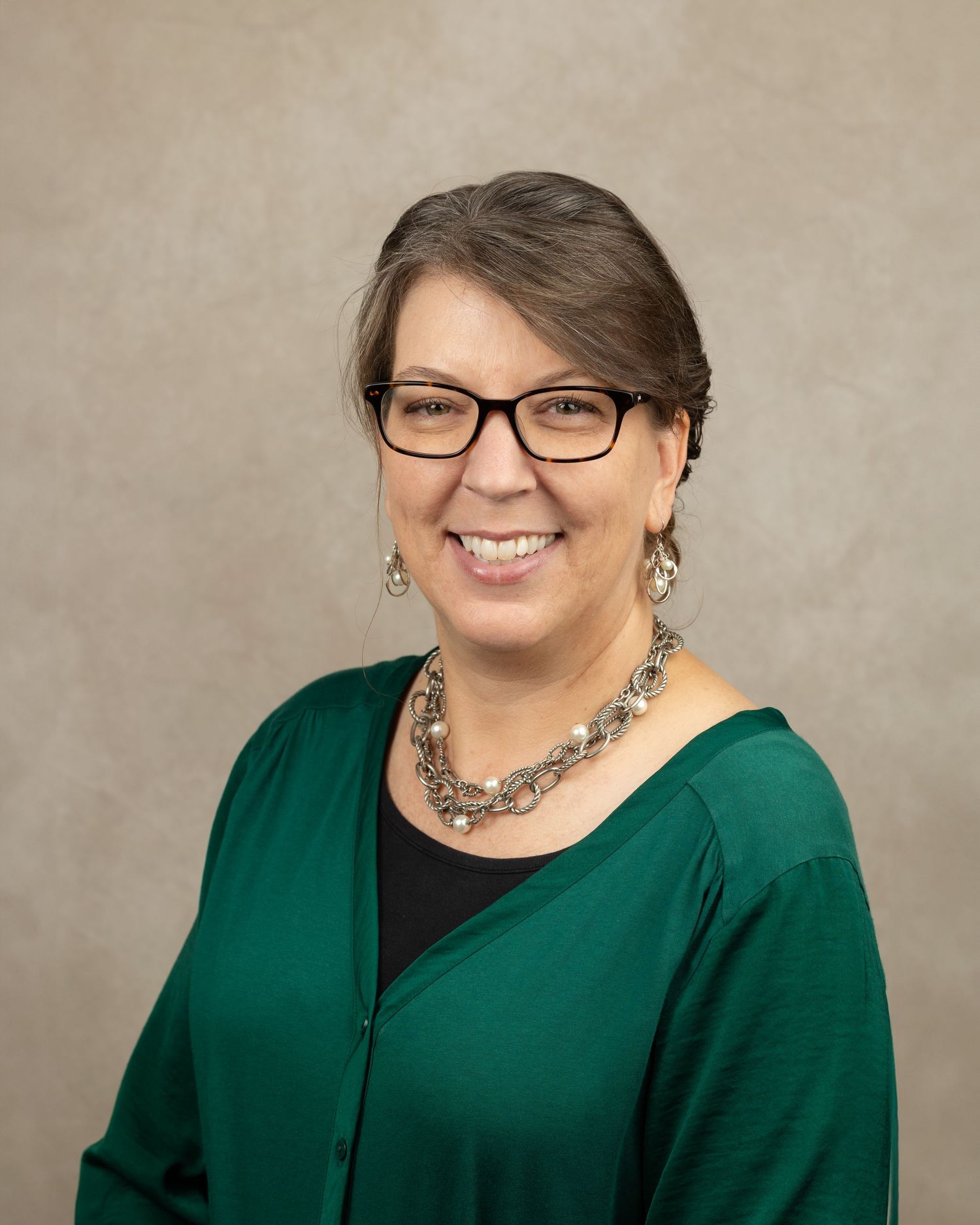 Studio portrait of a person in a green button-up cardigan with glasses and statement jewelry.