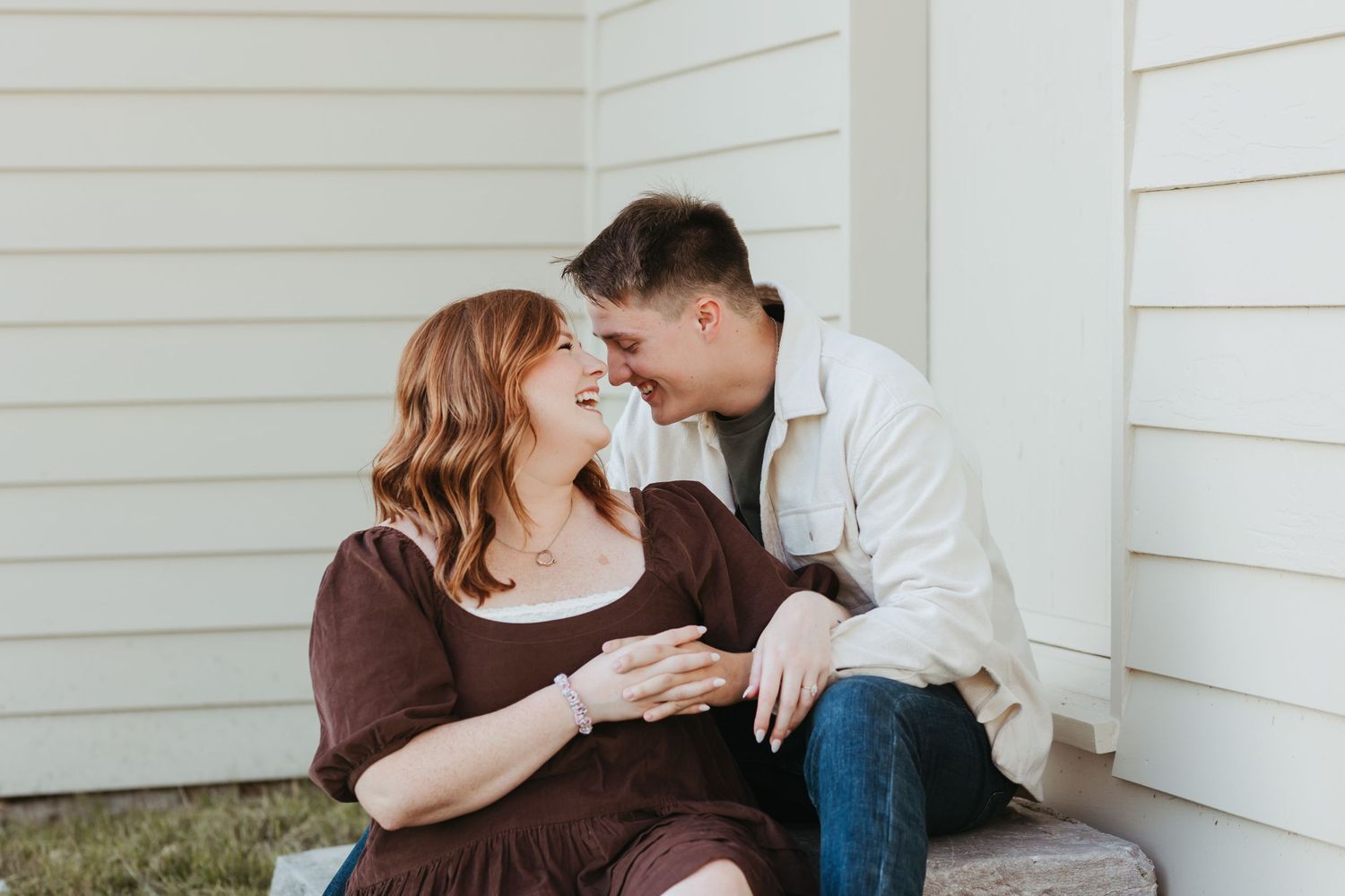 Couple sharing tender moment while sitting against white house exterior.