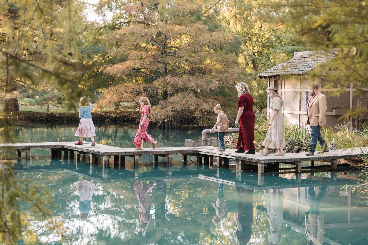 Group of people walking on wooden dock over reflective pond surrounded by autumn trees.