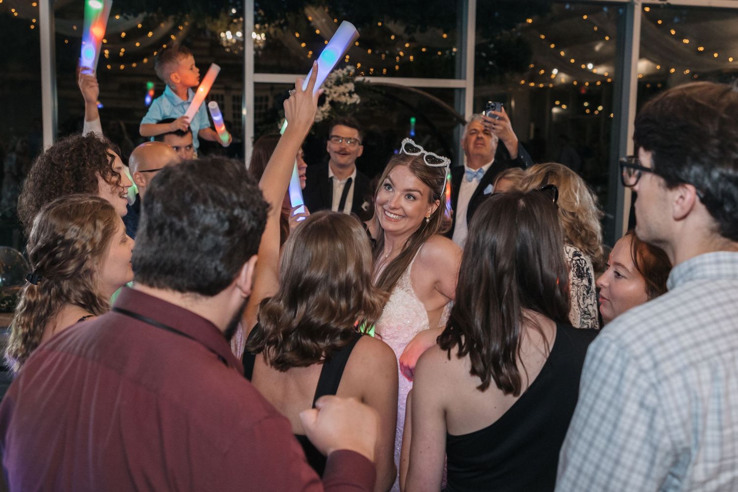 Wedding reception party guests dance with light-up batons under string lights indoors.