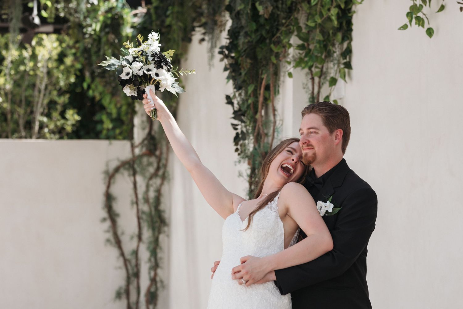 Joyful wedding couple celebrates with raised bouquet of white flowers.