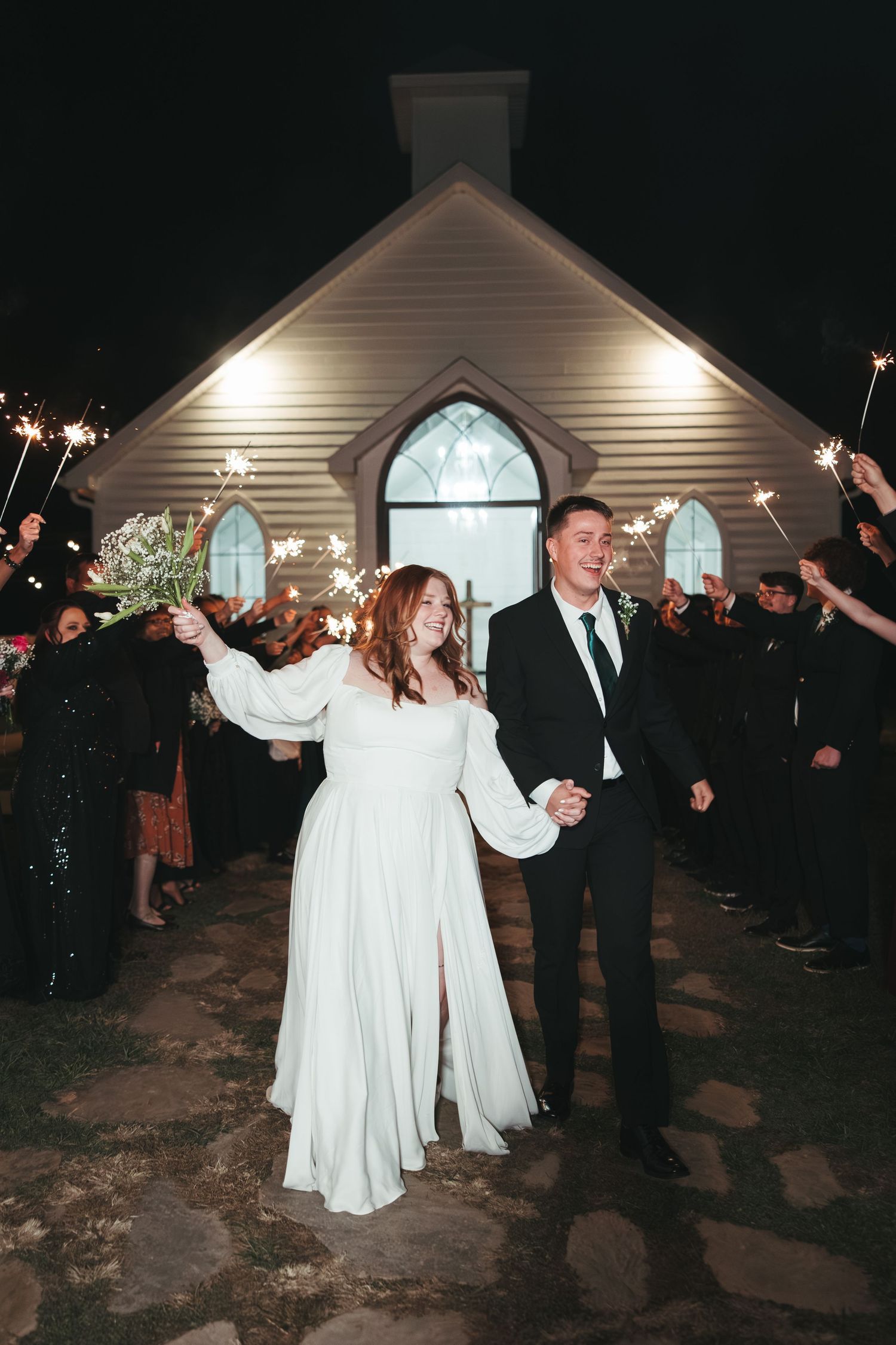 Wedding couple exits through sparkler tunnel outside illuminated chapel.