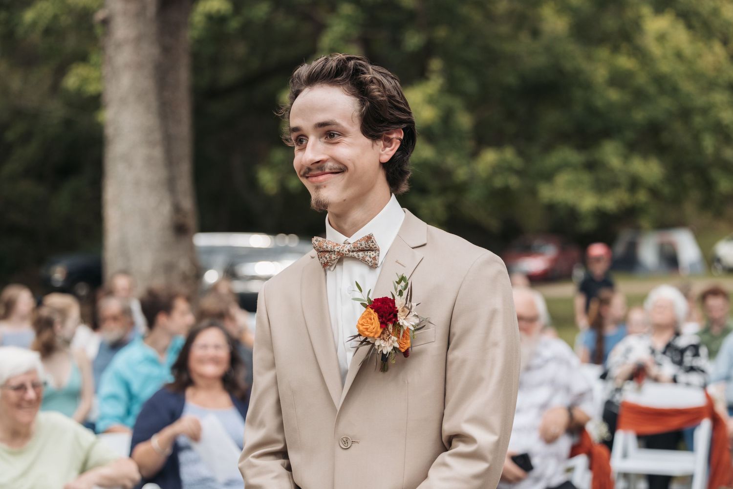 A member of the wedding party in a beige suit with orange boutonniere smiles during the ceremony.