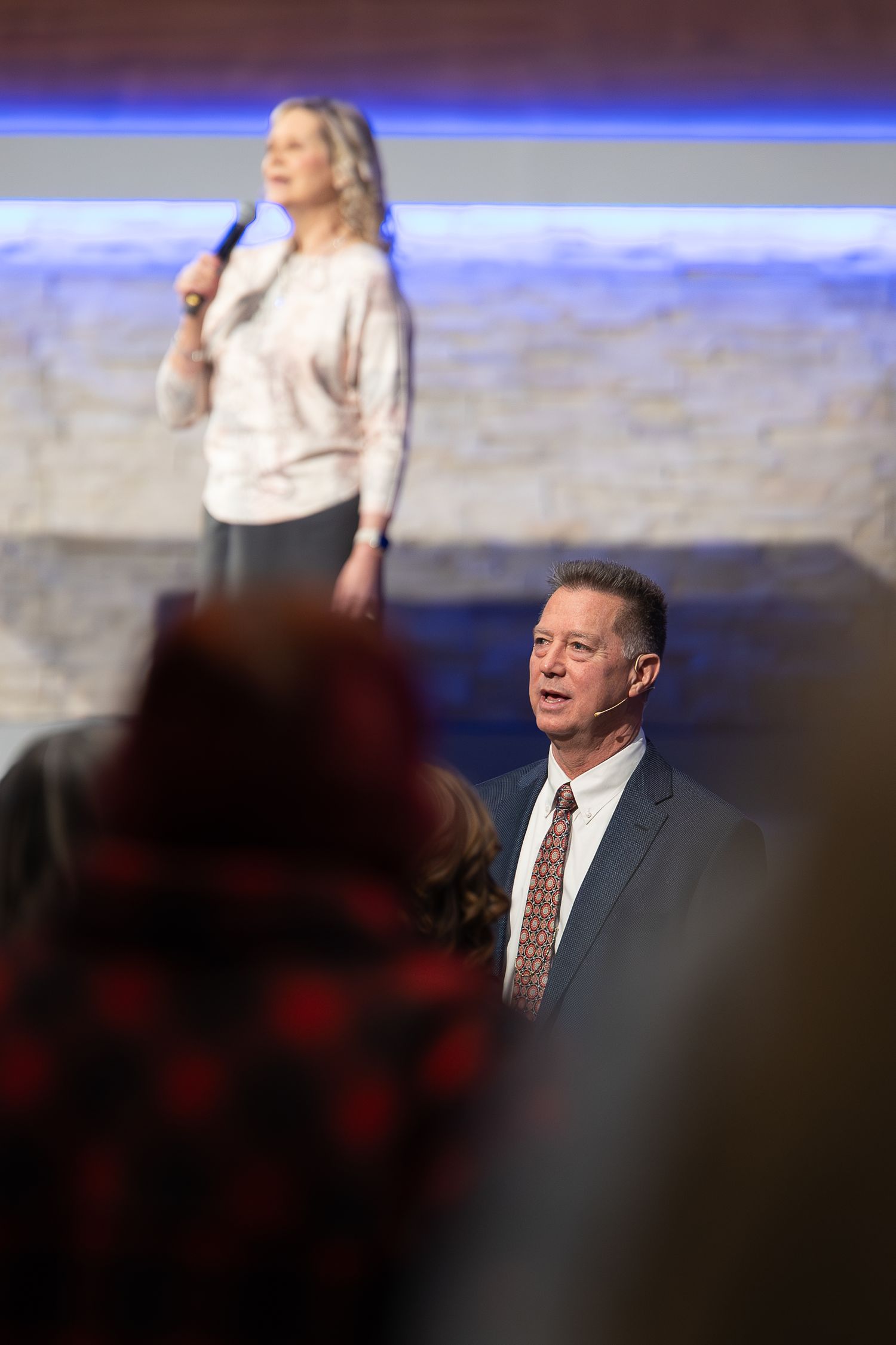 Conference speakers presenting on stage with dramatic blue lighting and audience in foreground.