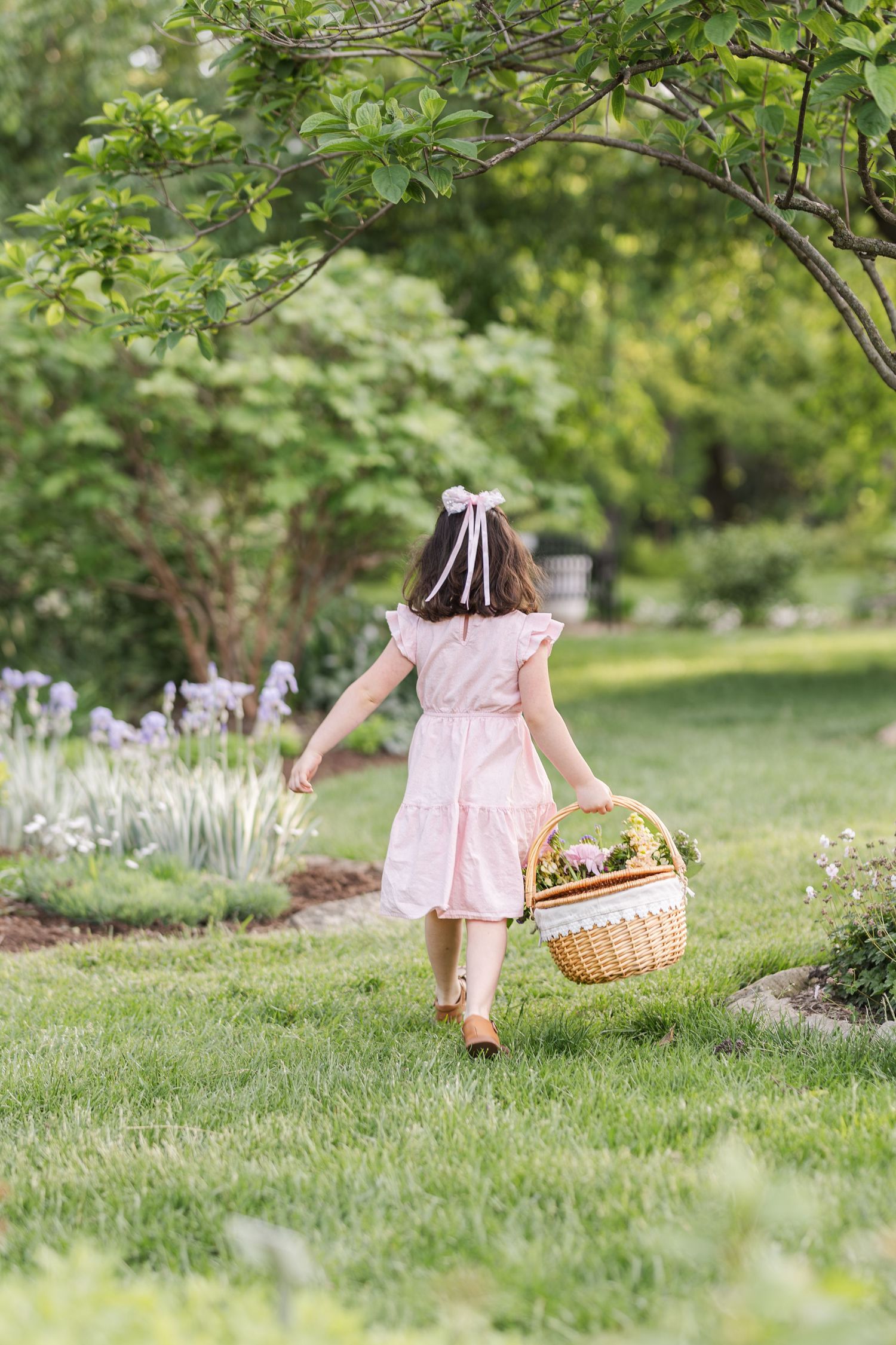 A pink-dressed figure walks through a garden path carrying a woven basket on a sunny summer day.