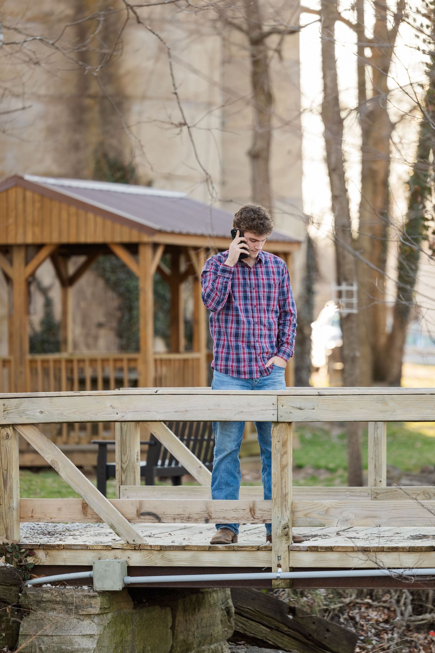 Person in plaid shirt and jeans standing on wooden bridge with gazebo in background.