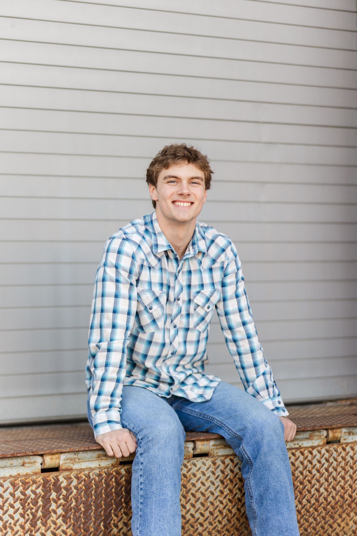 A smiling person in a blue and white plaid shirt and jeans sits casually on a wooden bench against a gray wall.