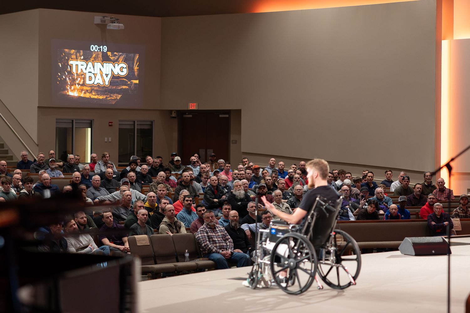A speaker in a wheelchair addresses a large audience seated in an auditorium with dim lighting.